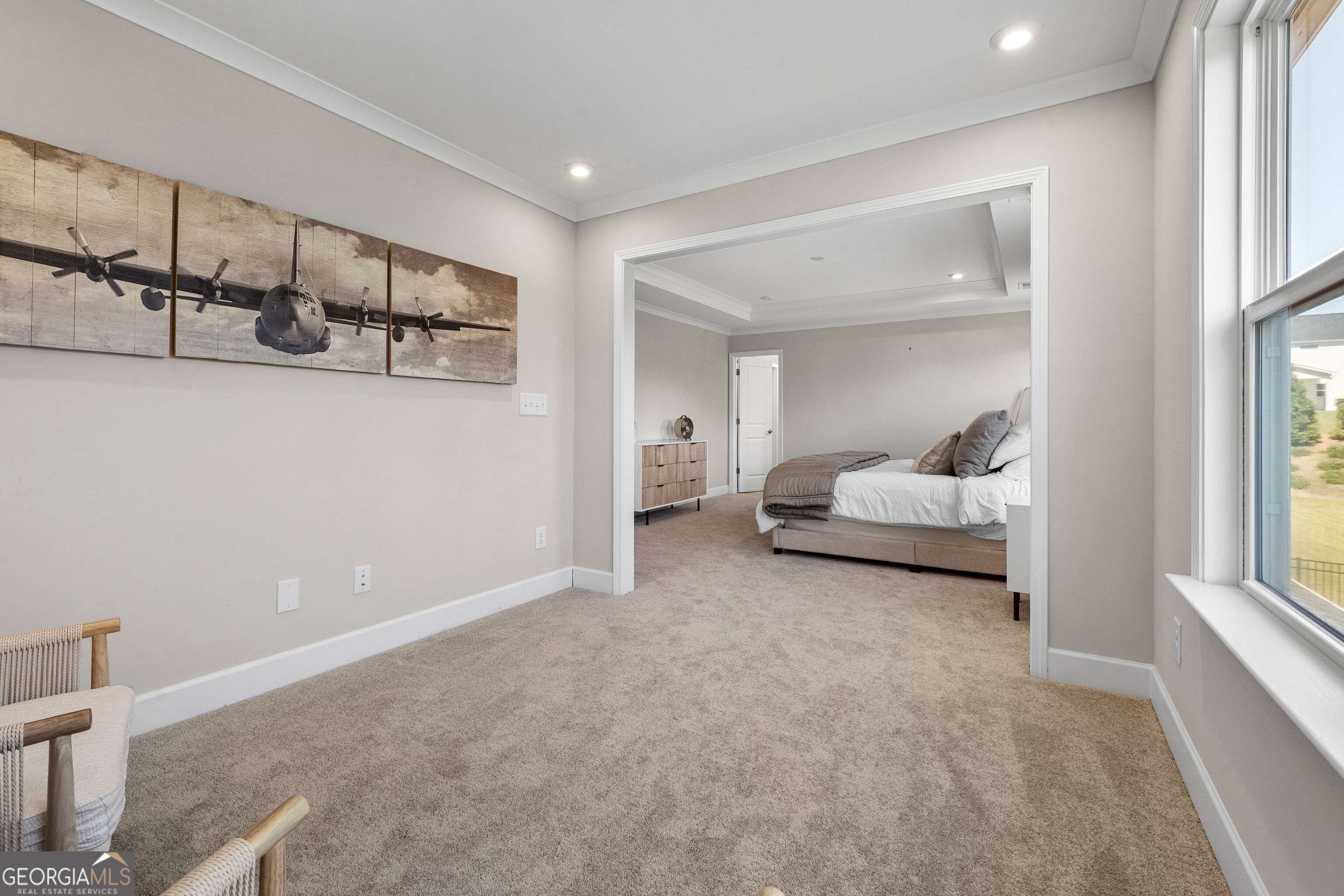 Carpeted sitting area opening into a bedroom with a bed, large windows, and airplane wall art.