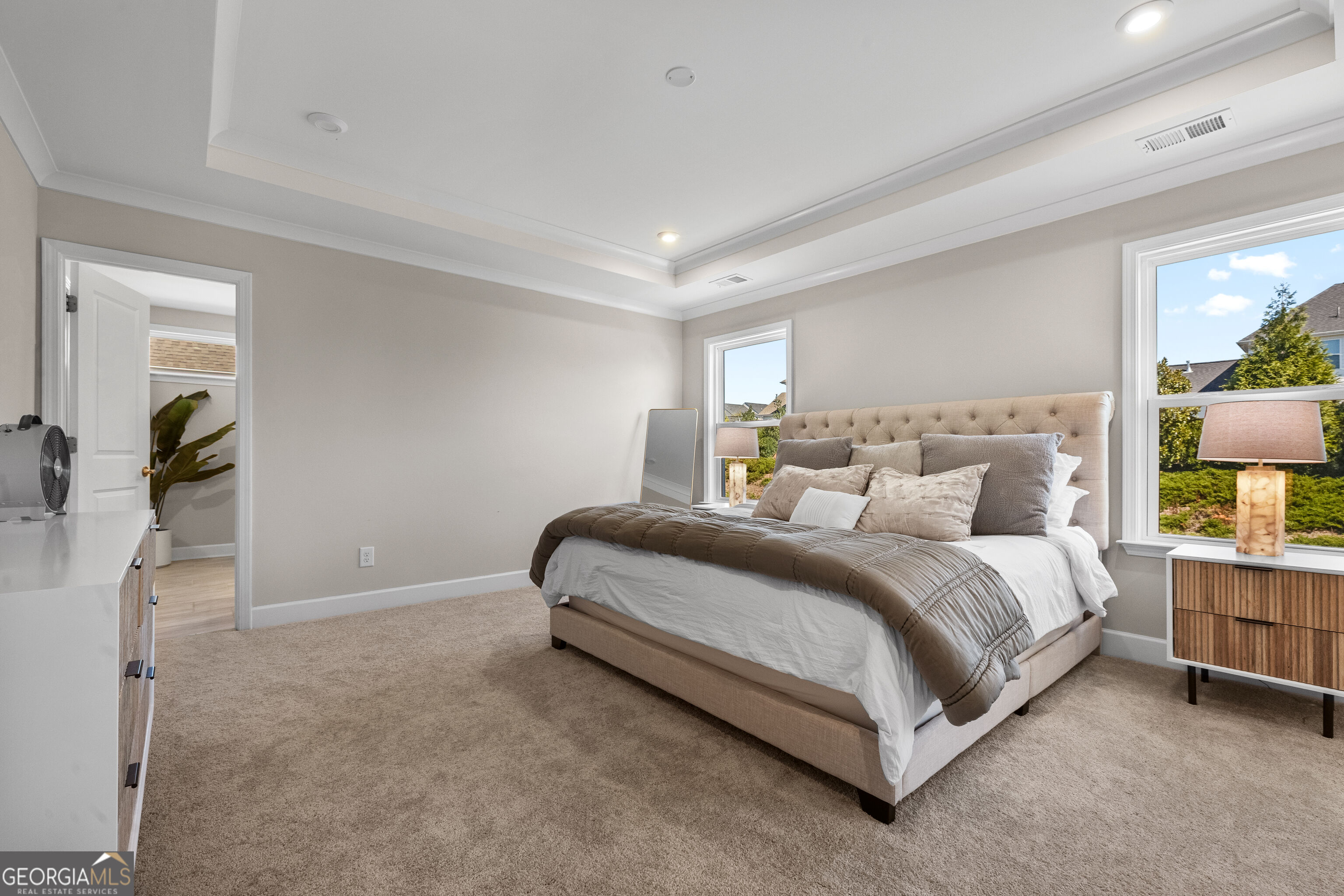 Furnished bedroom with a tufted bed, tray ceiling, beige carpet, and two large windows.