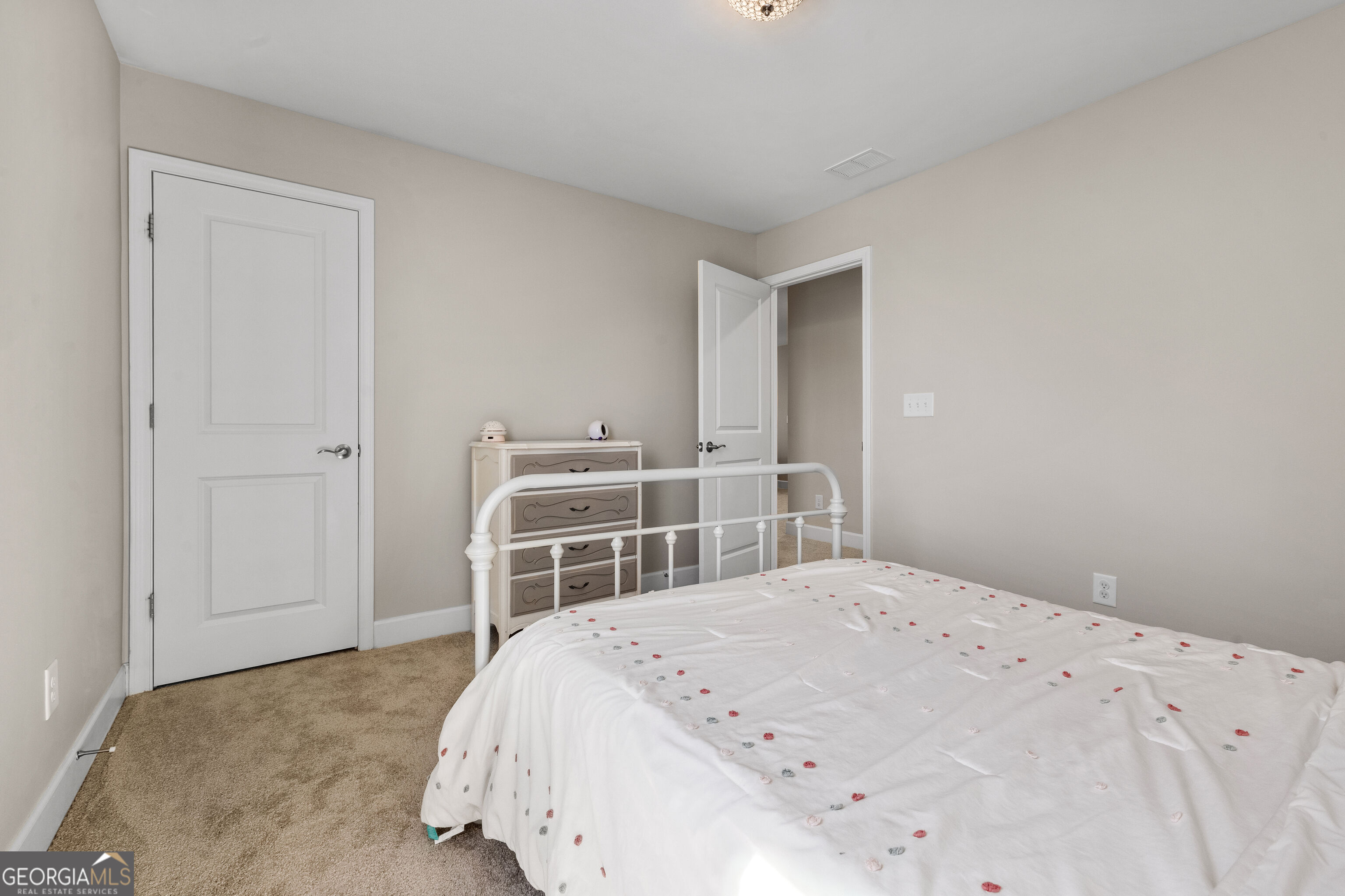 Bedroom with a white metal bed, patterned coverlet, beige carpet, and dresser.