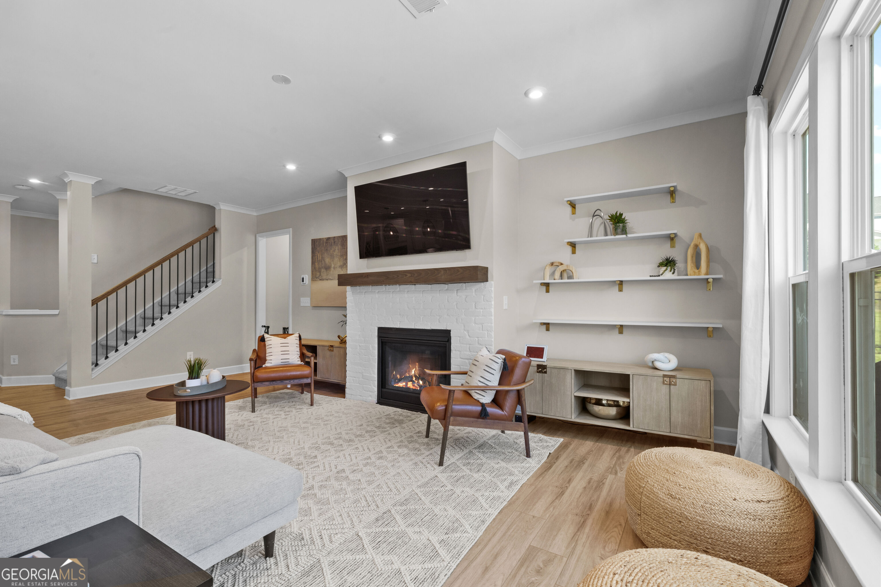 Staged living room with white brick fireplace, mounted TV, floating shelves, and large windows.