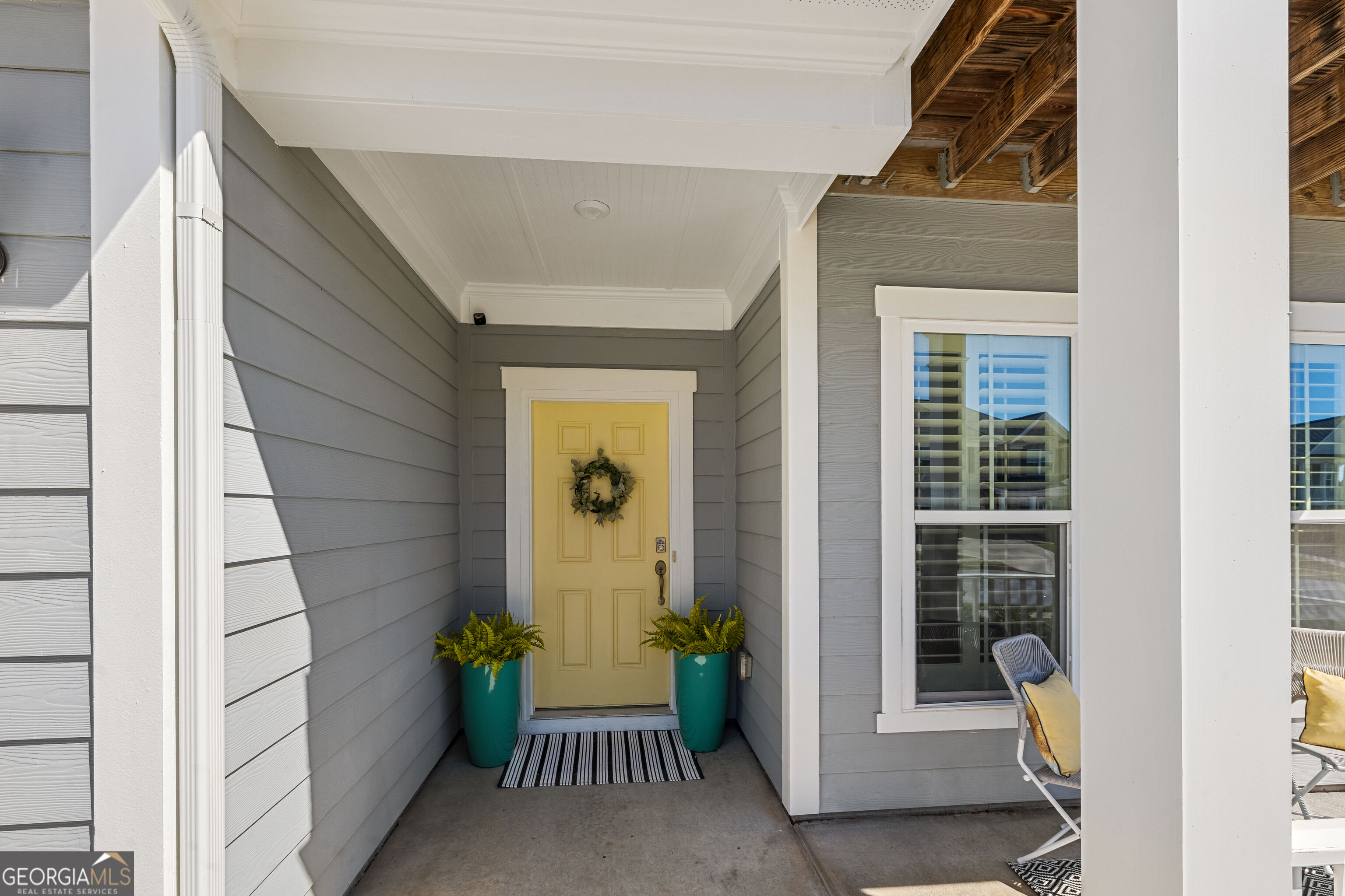 Covered porch with a yellow wreath-adorned door, turquoise fern planters, and exposed wood ceiling beams.