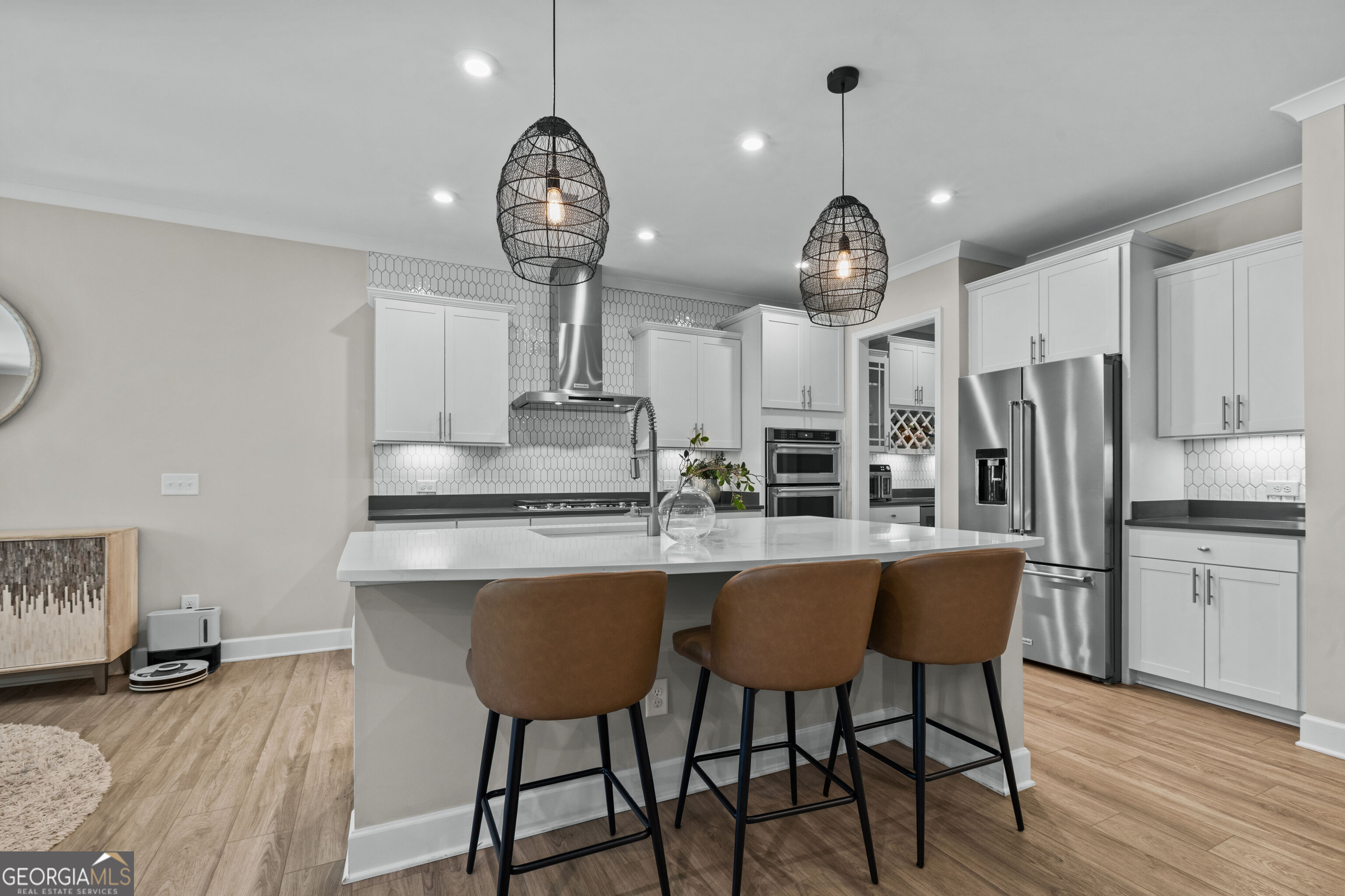 Modern kitchen with a white island, three brown stools, stainless steel appliances, and black pendant lights.