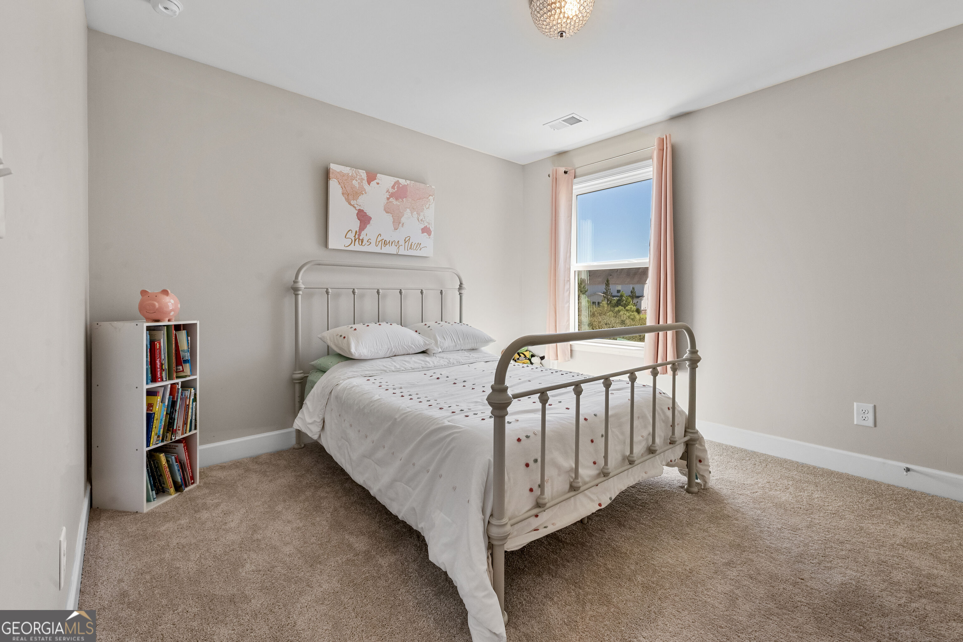 Bedroom with a white metal bed, pink curtains, and a bookshelf topped with a piggy bank.