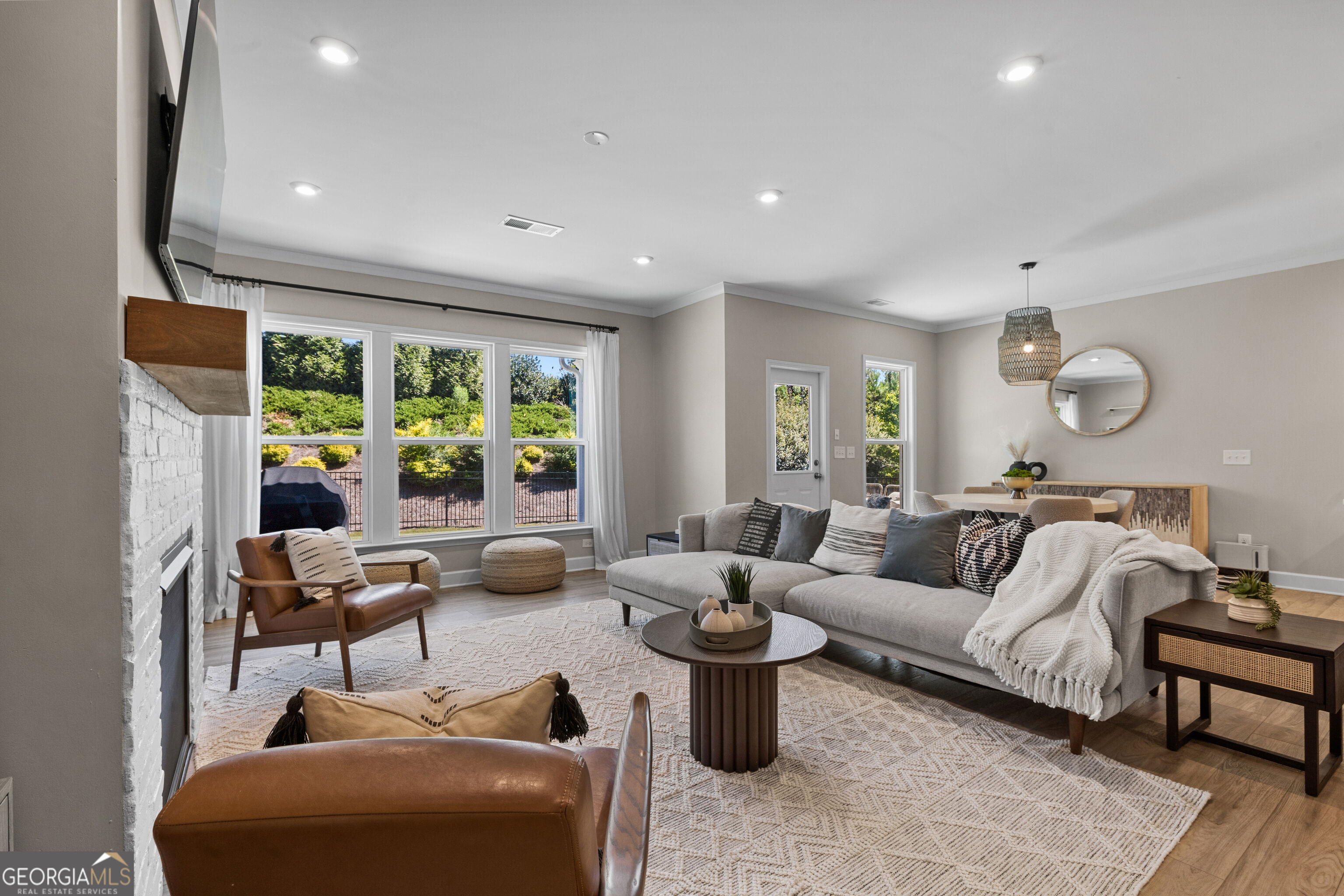 Staged living room with a gray sectional, white brick fireplace, and large windows overlooking greenery.