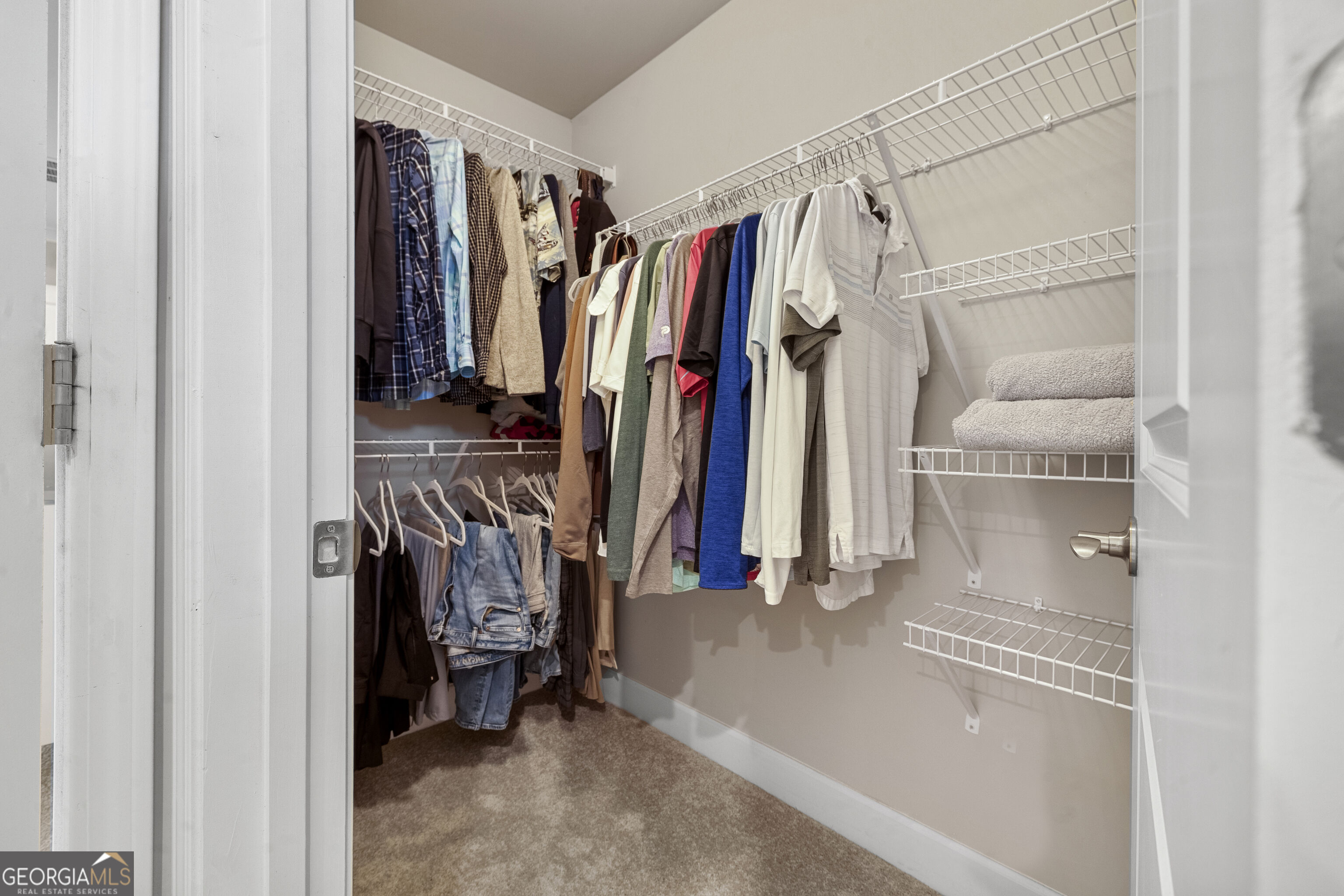 Walk-in closet with clothes on wire racks, empty shelves, folded towels, and carpeted flooring.