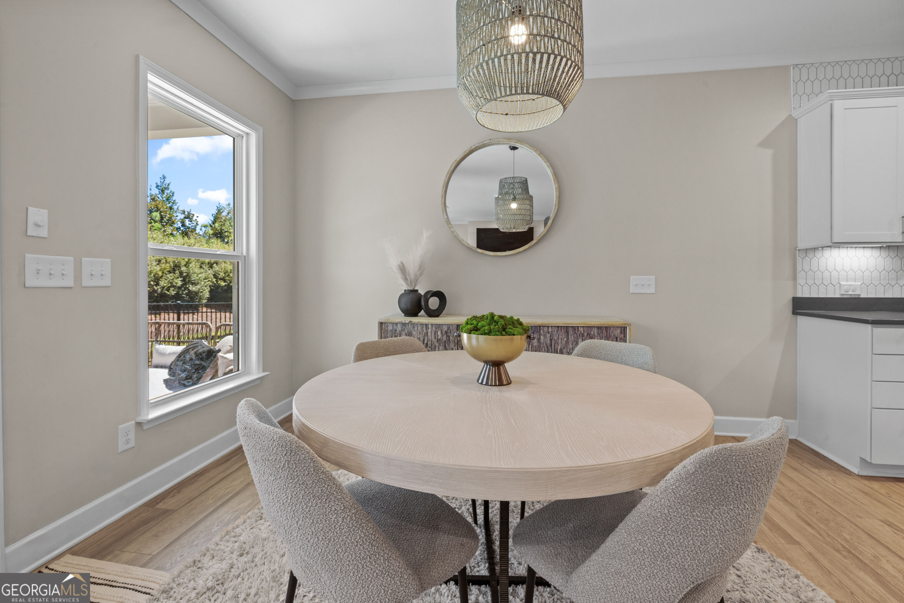 Dining area with a round table, textured chairs, woven pendant light, and large window overlooking trees.