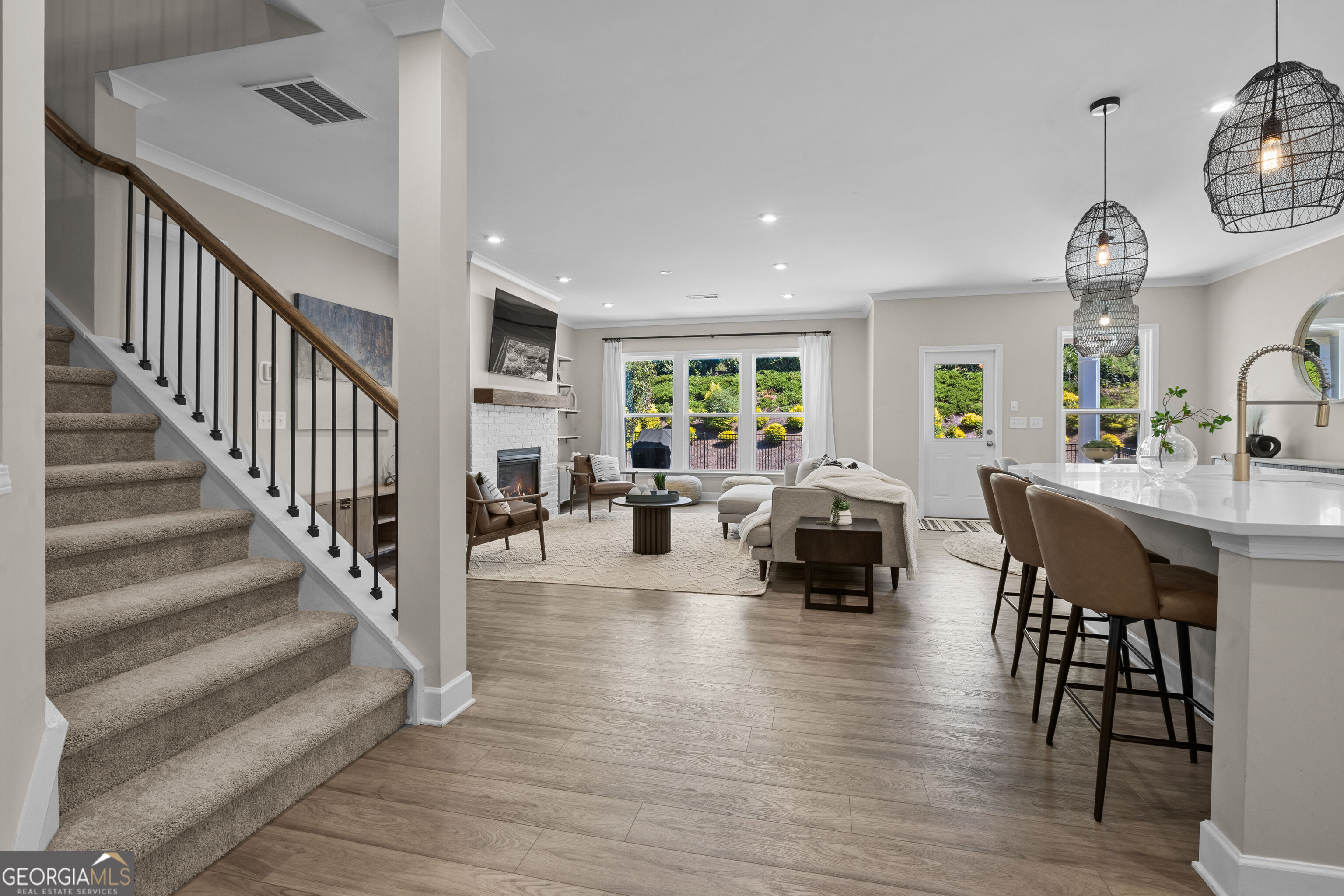 Open-plan living room and kitchen with a carpeted staircase, white brick fireplace, and island pendant lights.