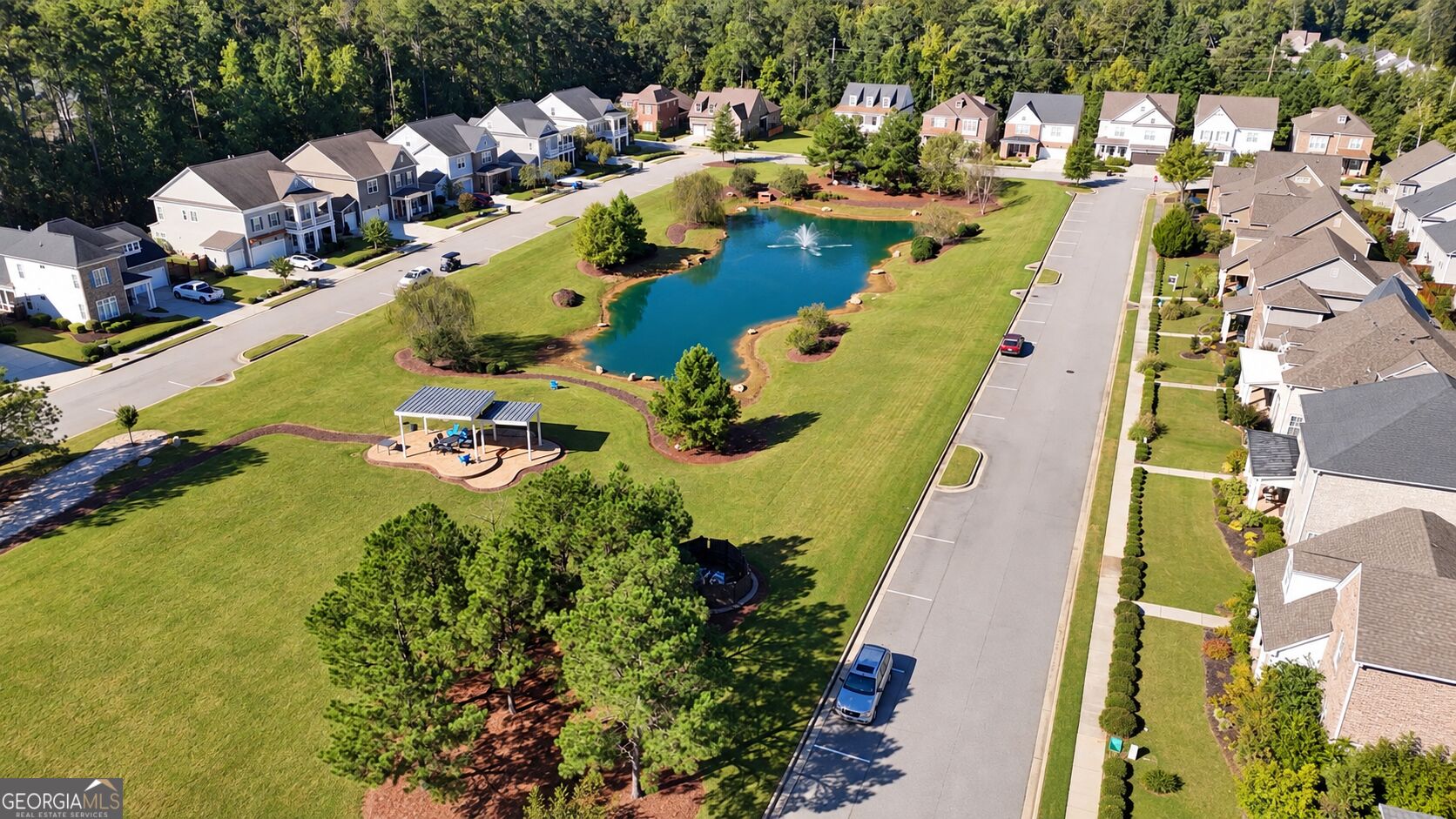 Aerial view of a neighborhood pond with a fountain and covered pavilion, surrounded by homes and lawns.