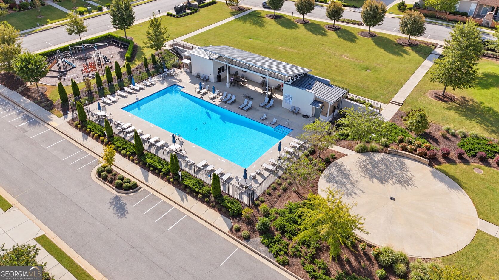 Aerial view of a community pool with a covered pavilion, fenced deck, and nearby playground.