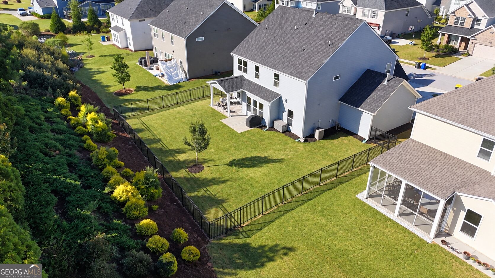 Aerial view of a fenced backyard with a broad lawn, covered patio, and landscaped garden beds.