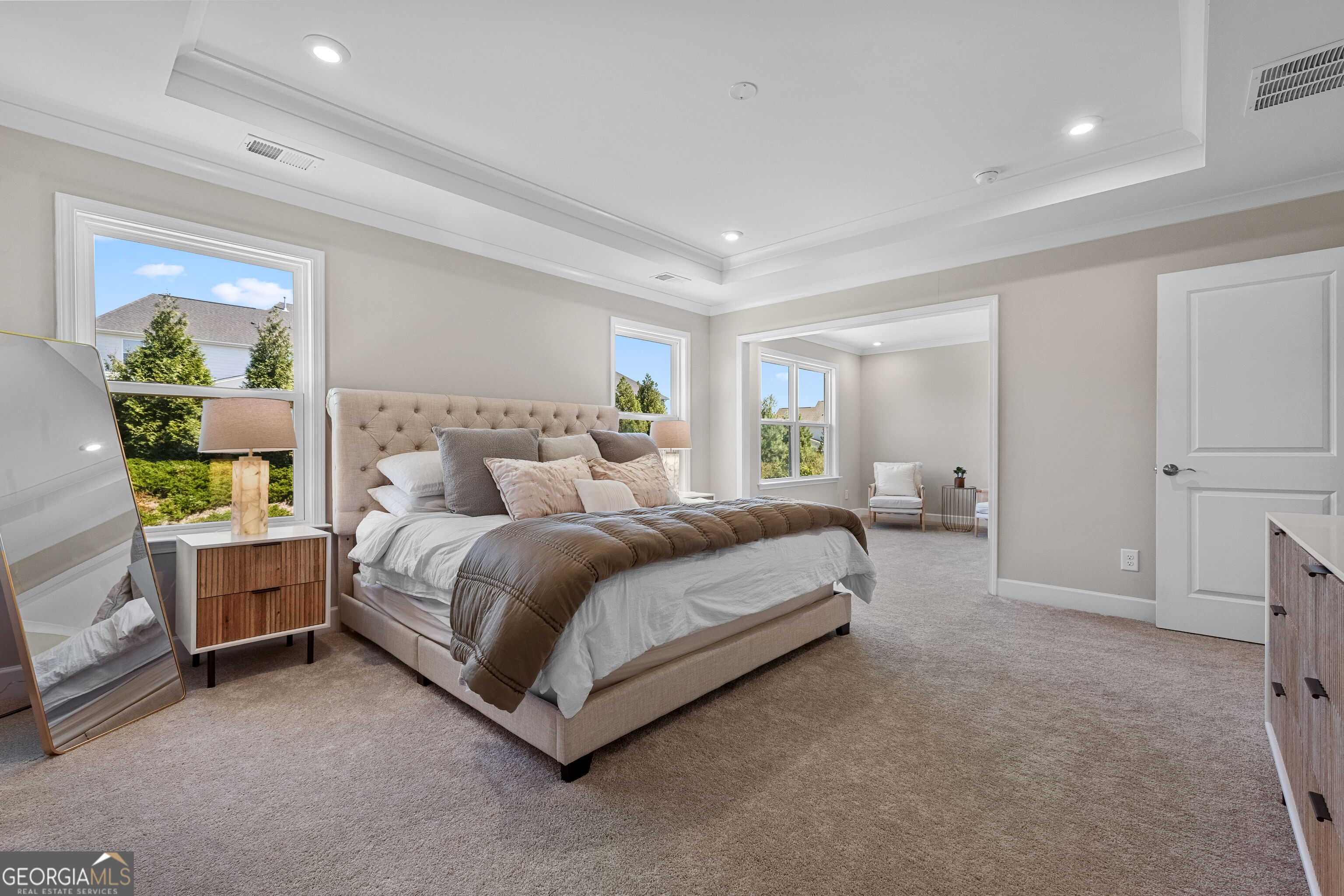 Staged bedroom with a tufted upholstered bed, carpet, and doorway to a bright sitting area.