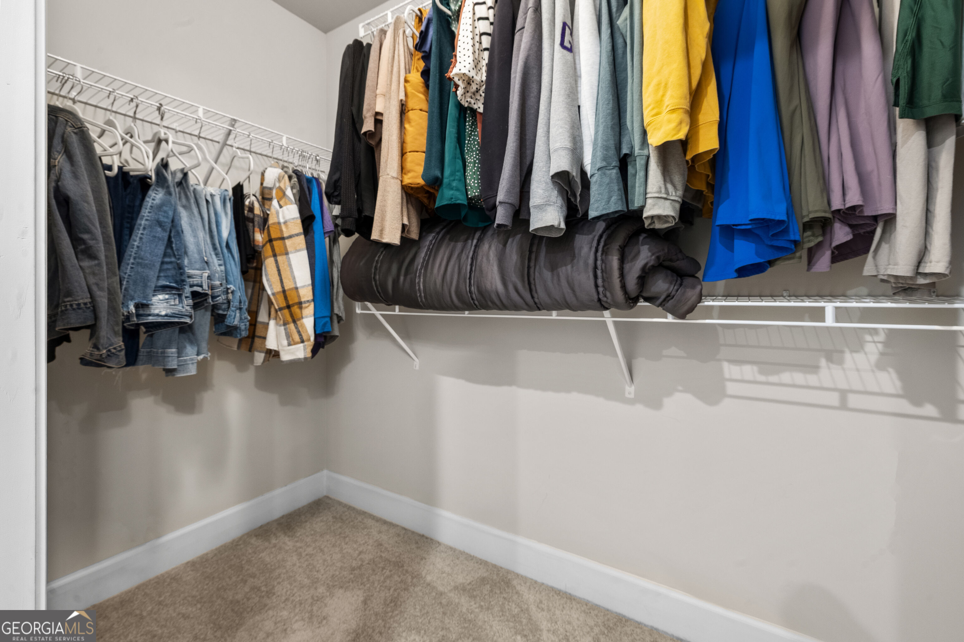 Walk-in closet with colorful hanging clothes, wire shelving, a folded comforter, and beige carpet.