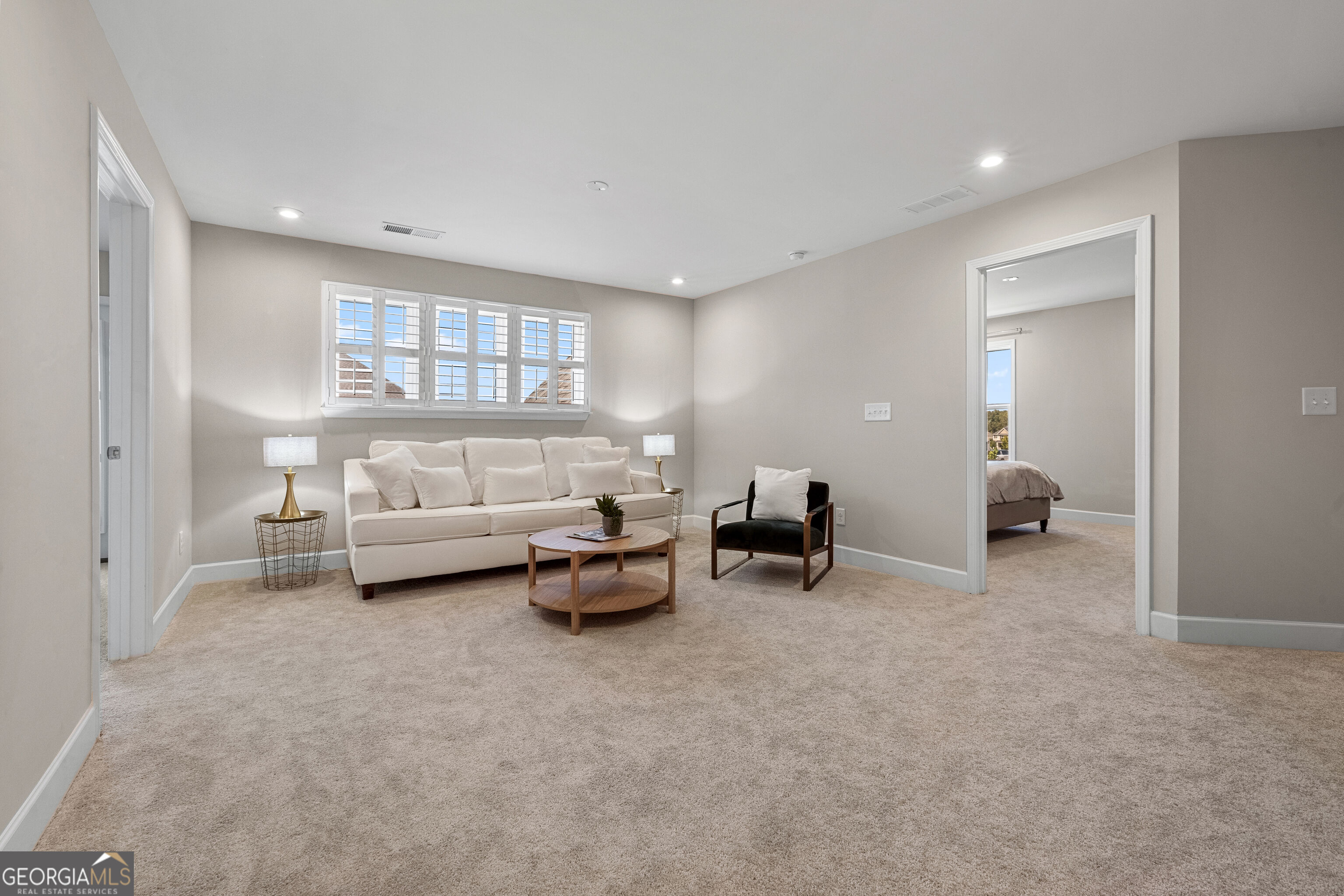 Living room with a white sofa beneath plantation-shuttered windows and an open doorway to a bedroom.