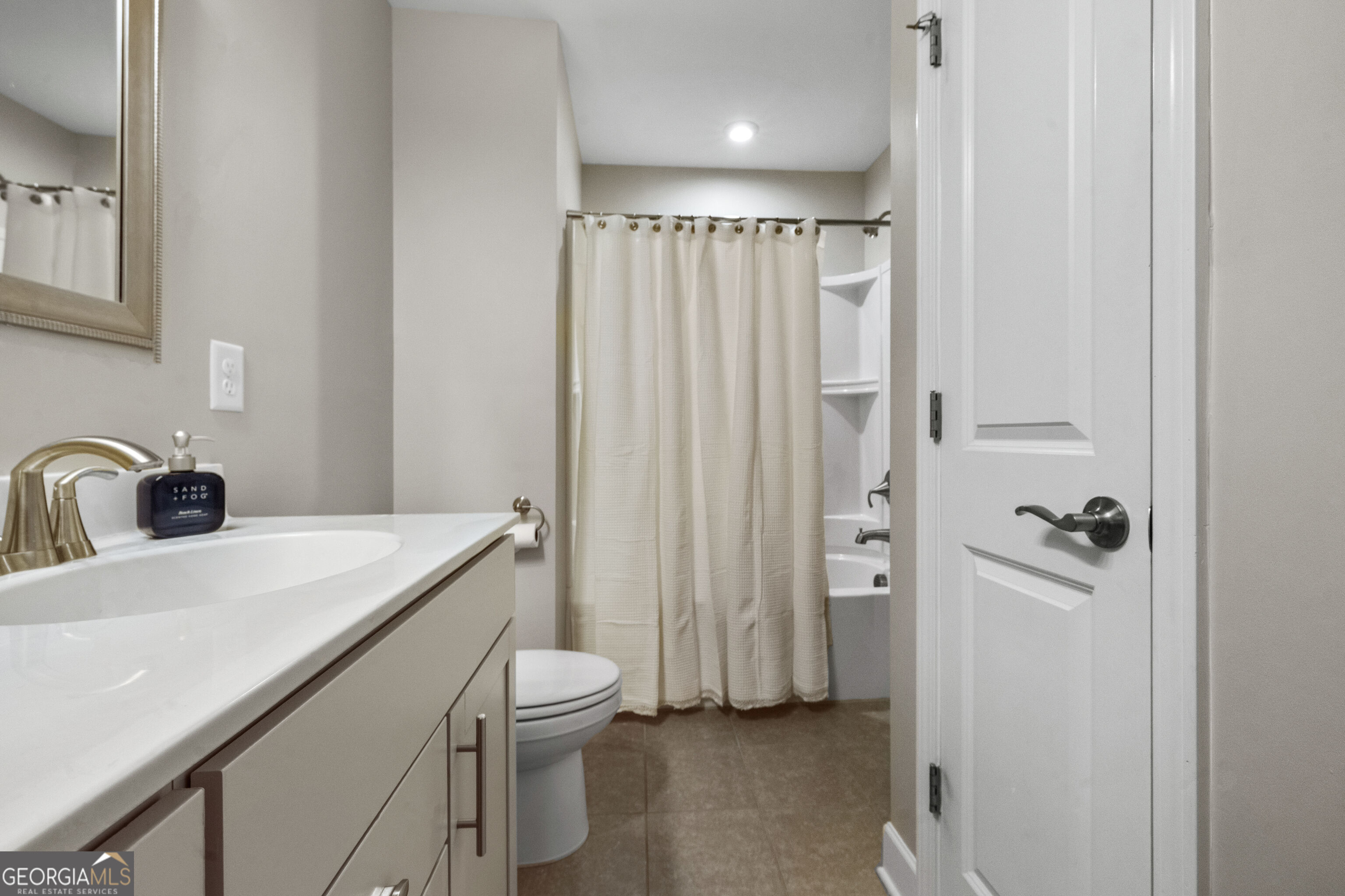 Bathroom with a white vanity sink, toilet, and cream shower curtain over brown tile flooring.