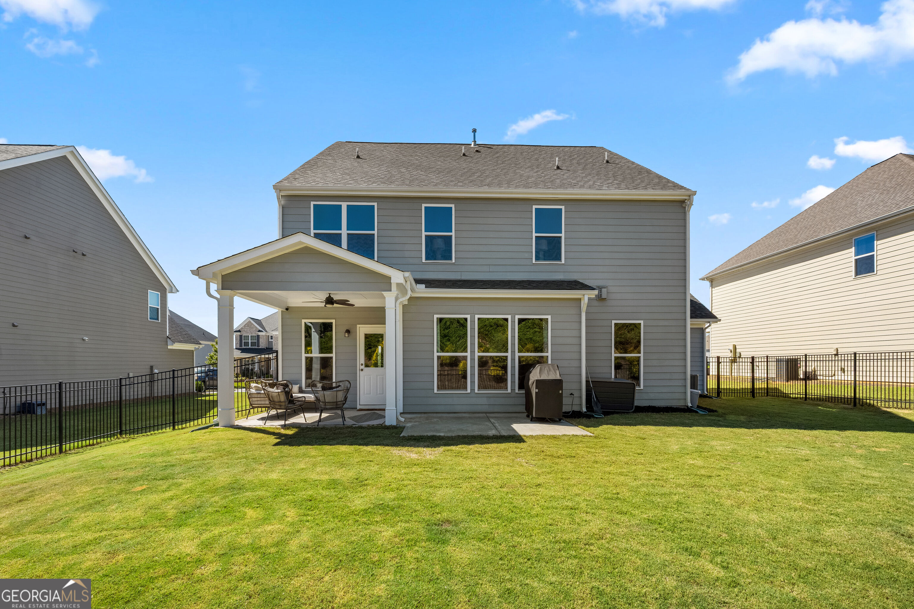 Rear of a two-story gray house with a covered patio, outdoor seating, and fenced lawn.