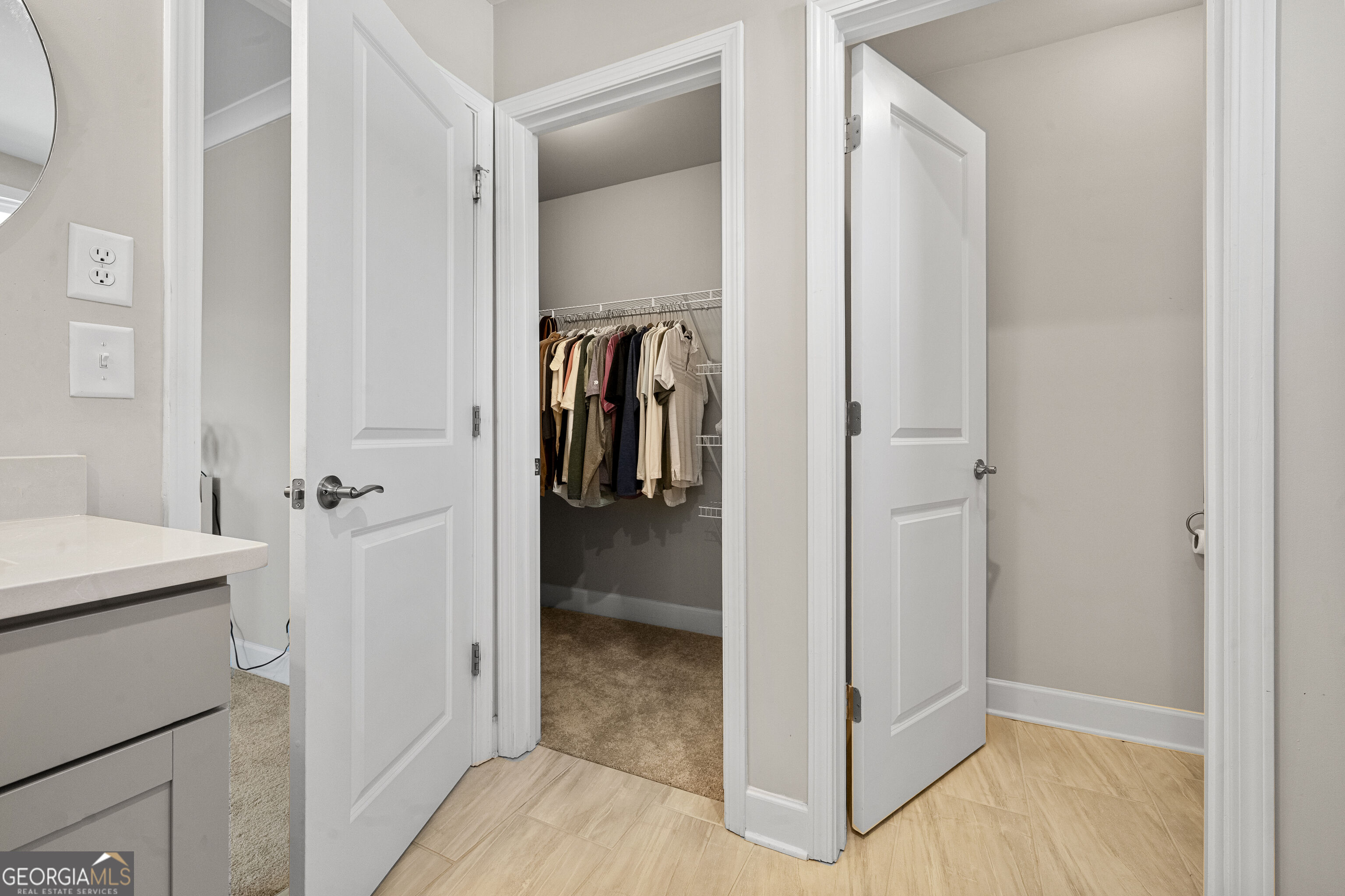Bathroom with a white vanity and tiled floor, opening to a walk-in closet with hanging clothes.