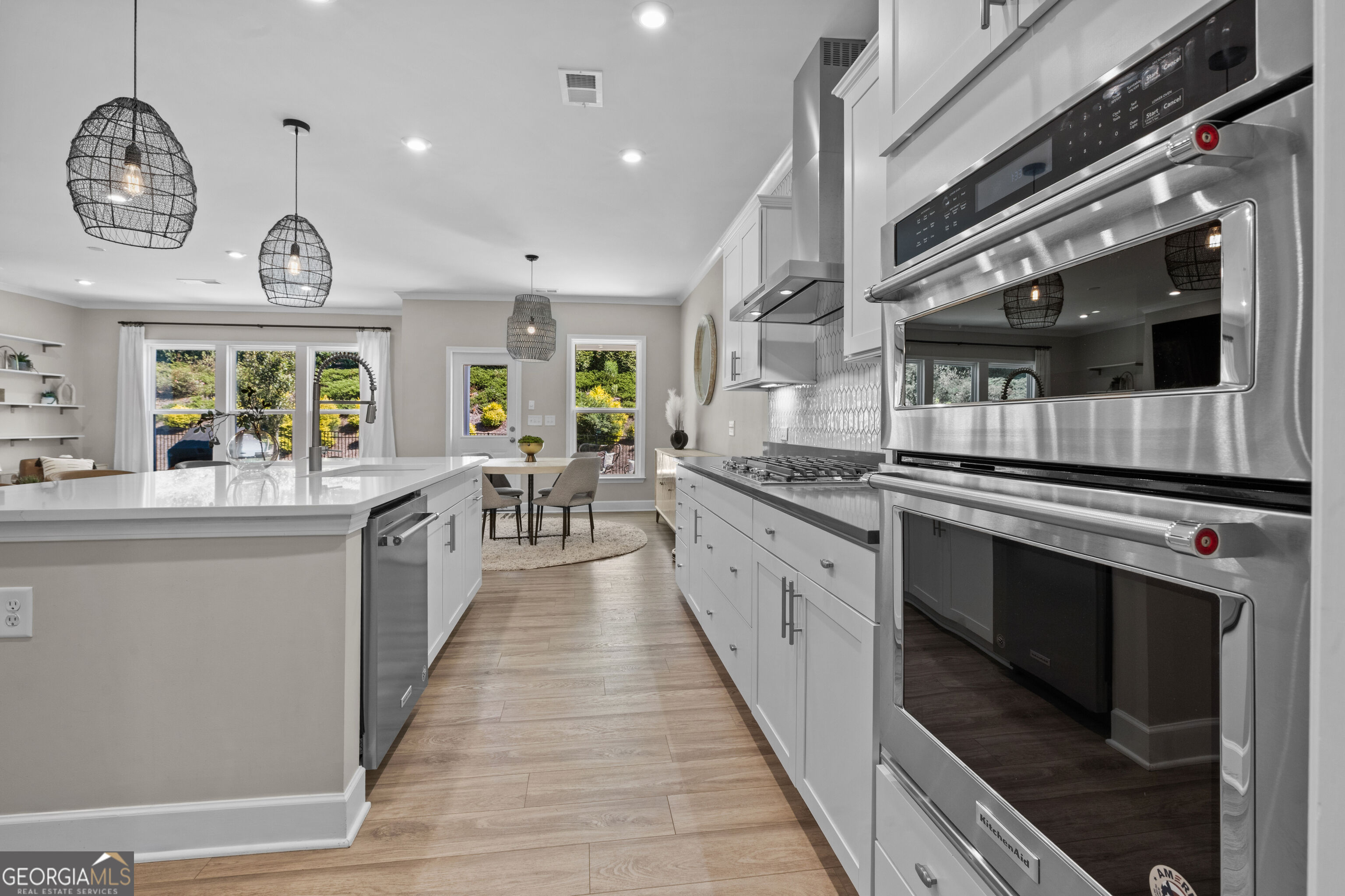 Modern white kitchen with a large island, stainless steel double ovens, and a dining area overlooking greenery.