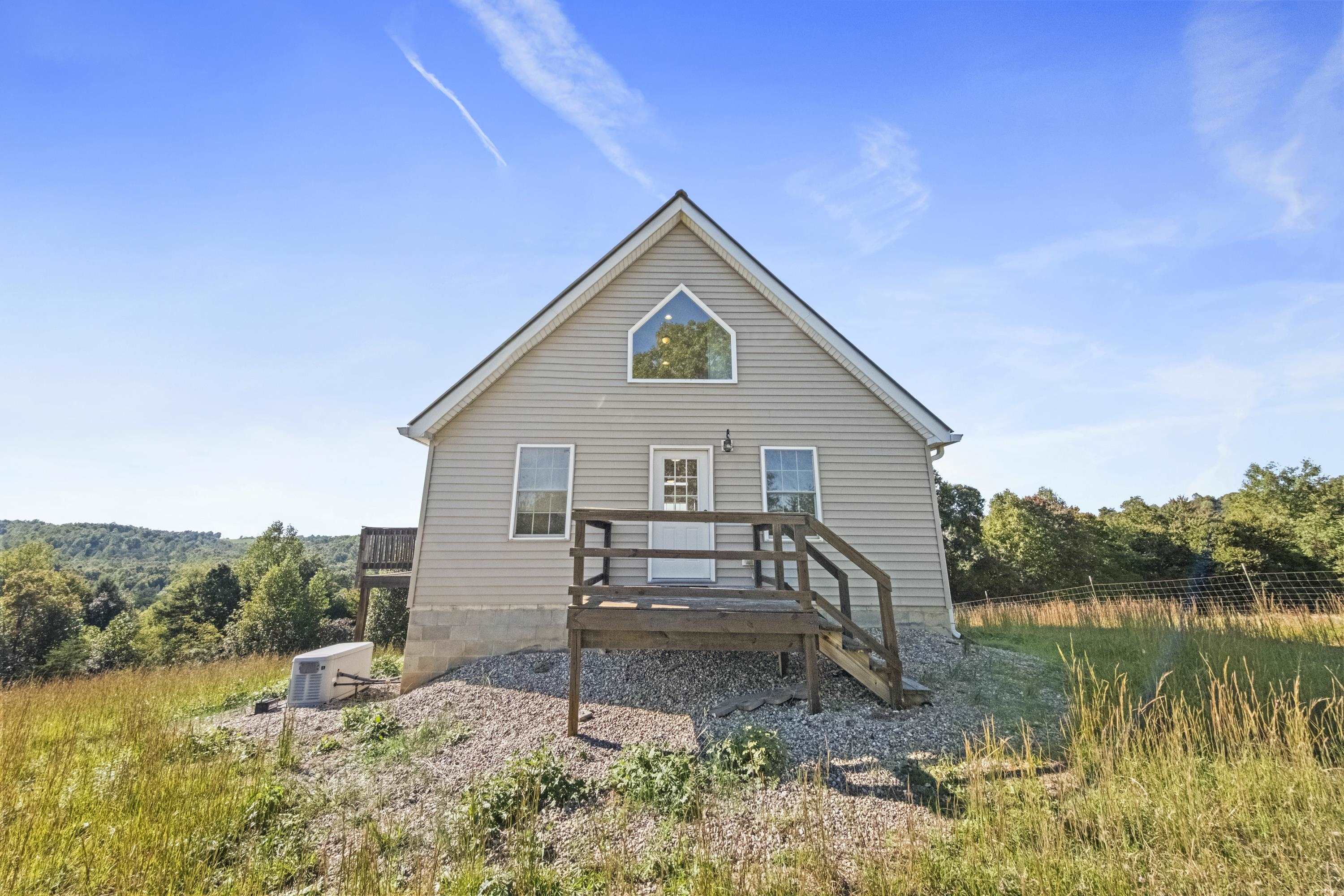 Gray-sided house with a steep gable, raised wooden deck, and wooded hills behind it.