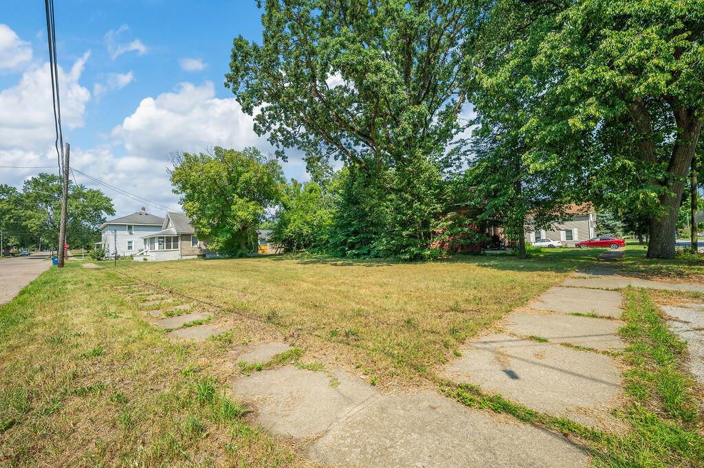 Vacant grassy lot with mature trees and a sidewalk along the street.