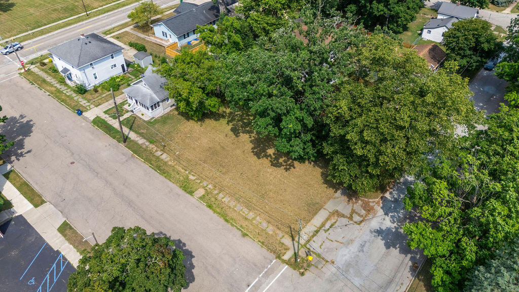 Aerial view of a vacant, tree-lined lot beside a street and neighboring homes.