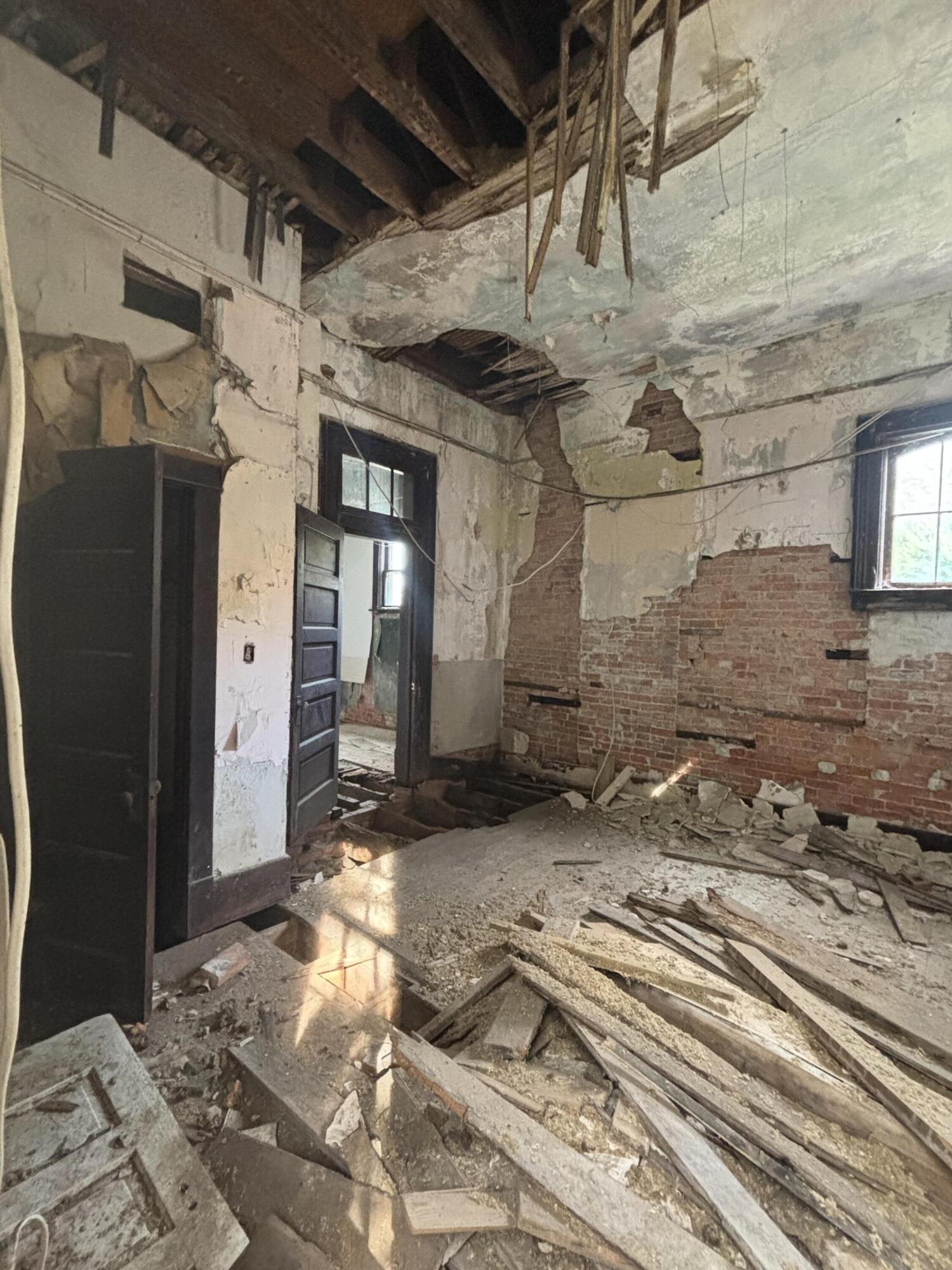 Gutted interior room with exposed brick, broken floorboards, debris, and missing sections of ceiling.