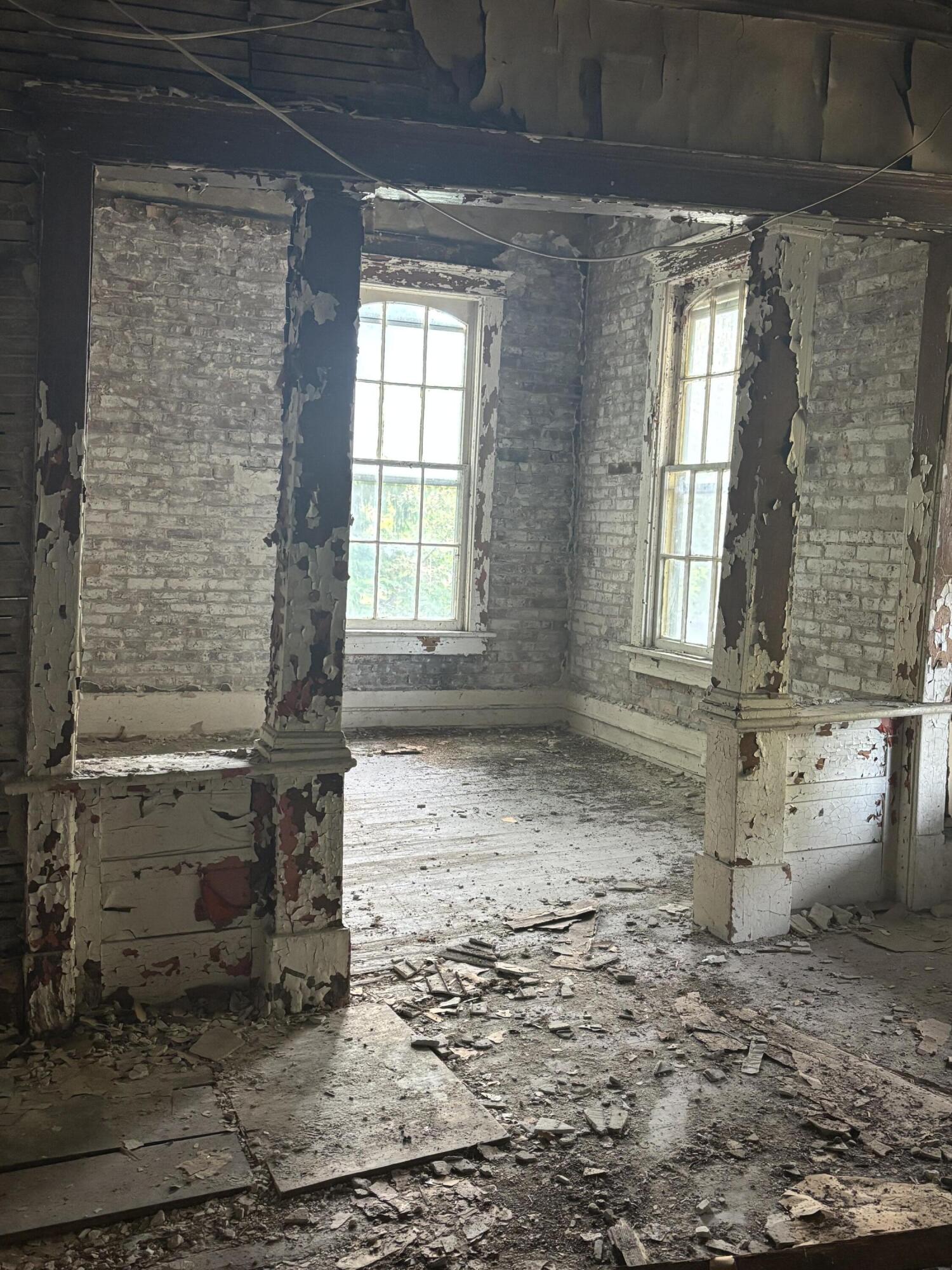 Vacant room with exposed brick, peeling paint, tall windows, and debris across the floor.