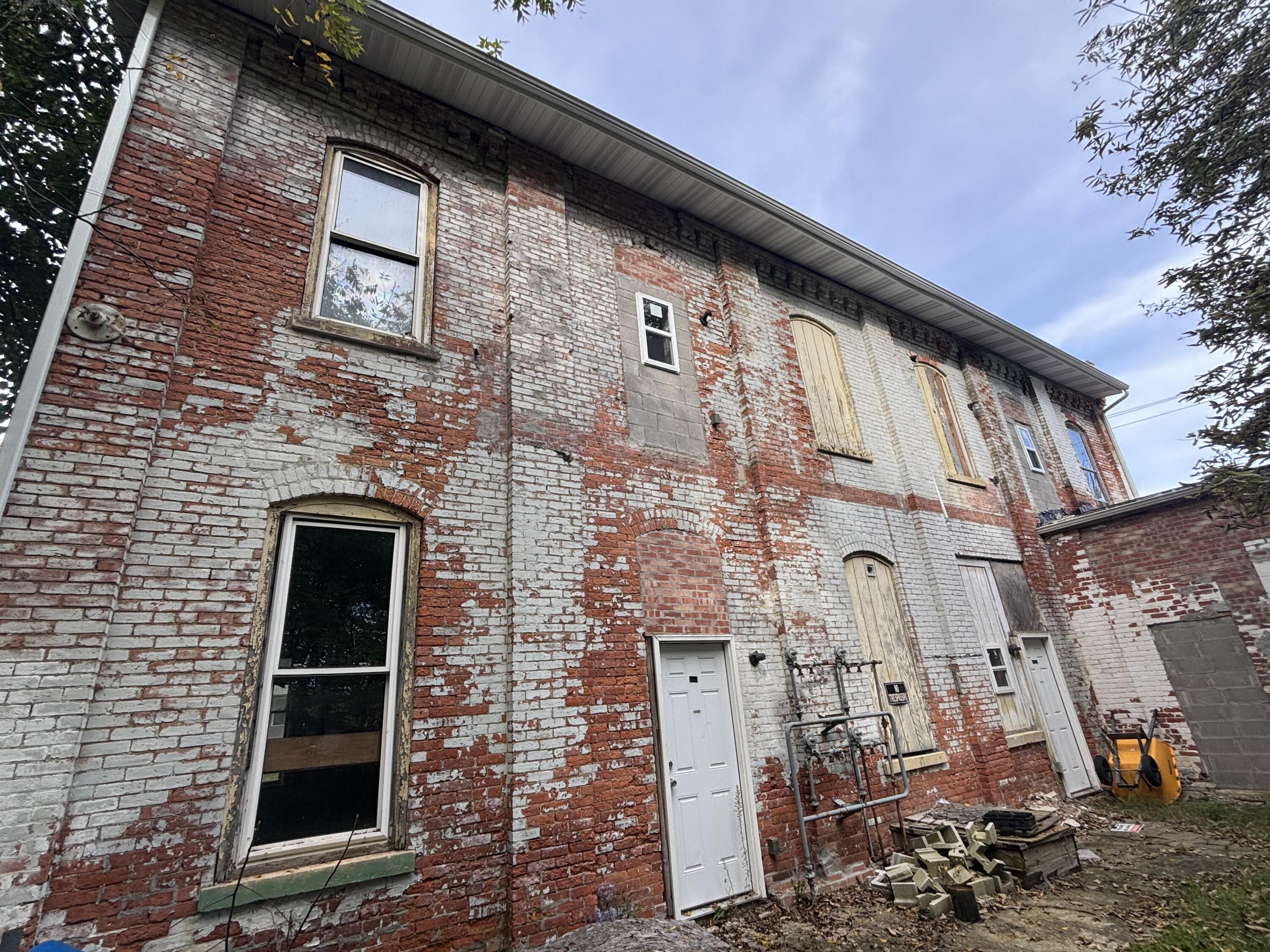 Two-story brick building with weathered white and red brick, boarded openings, exposed pipes, and debris.
