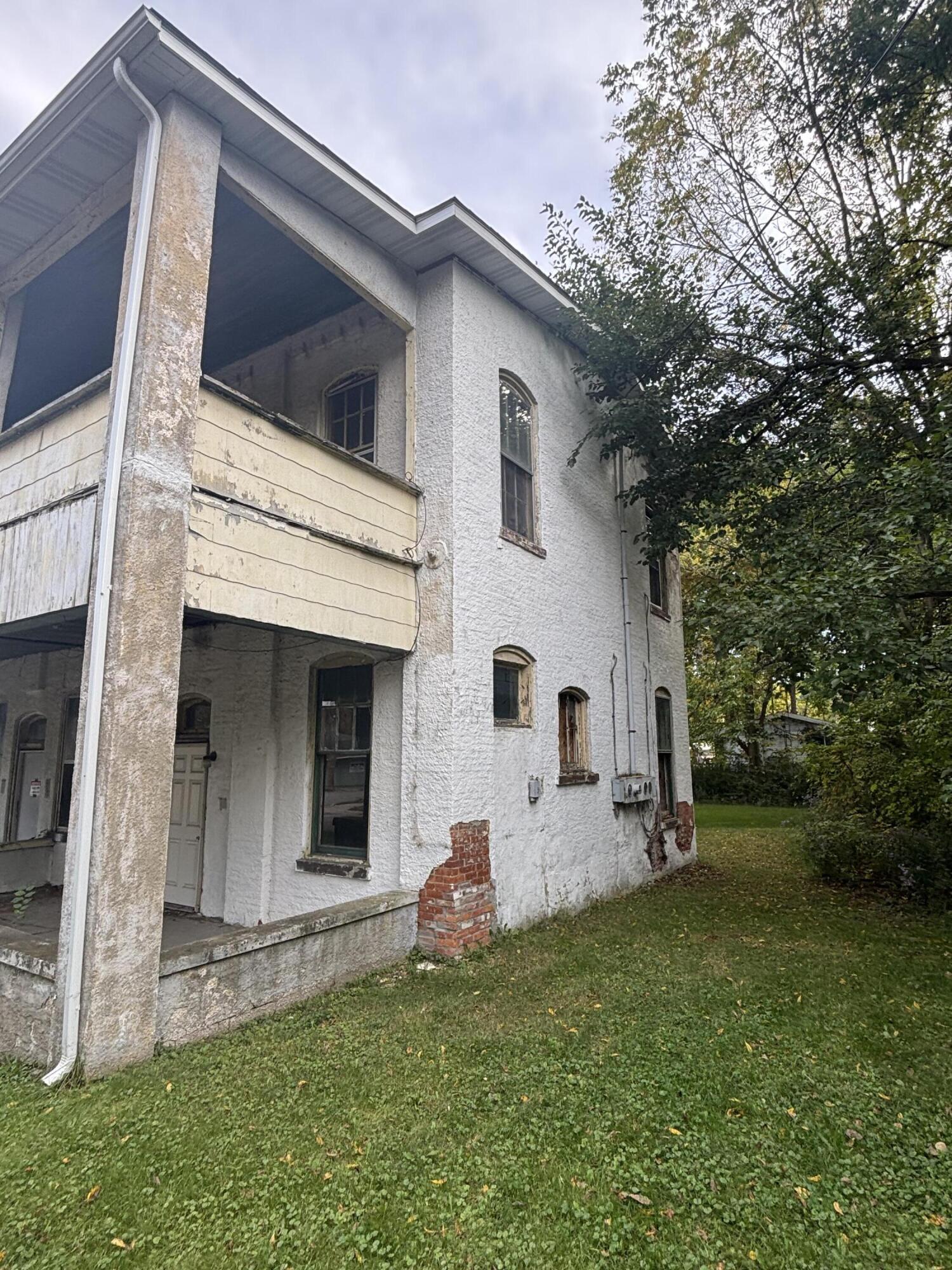 A weathered white house with a covered balcony, peeling siding, and exposed brick beside a leafy yard.