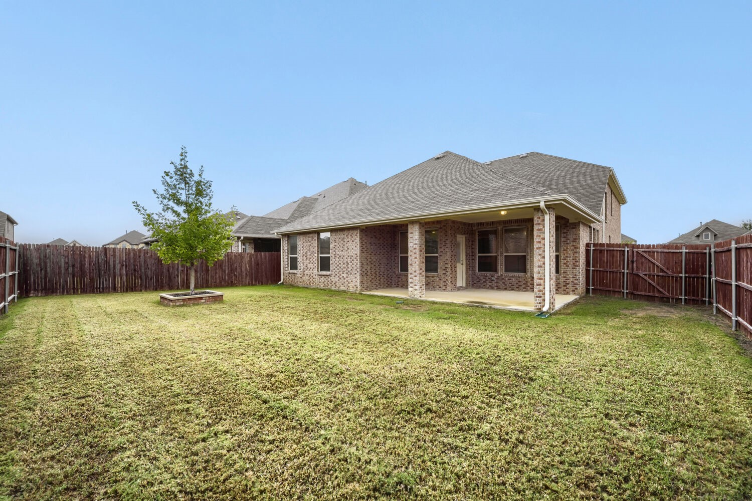 Brick home rear exterior with a covered patio, fenced lawn, and young tree.