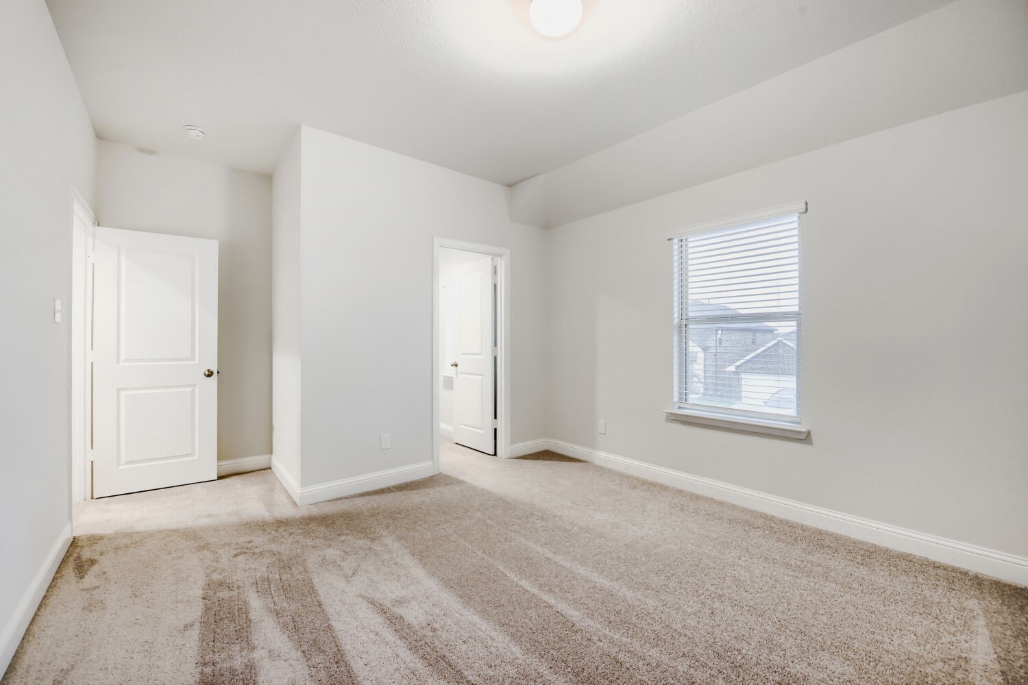 Empty carpeted bedroom with white walls, two open doors, and a window with blinds.