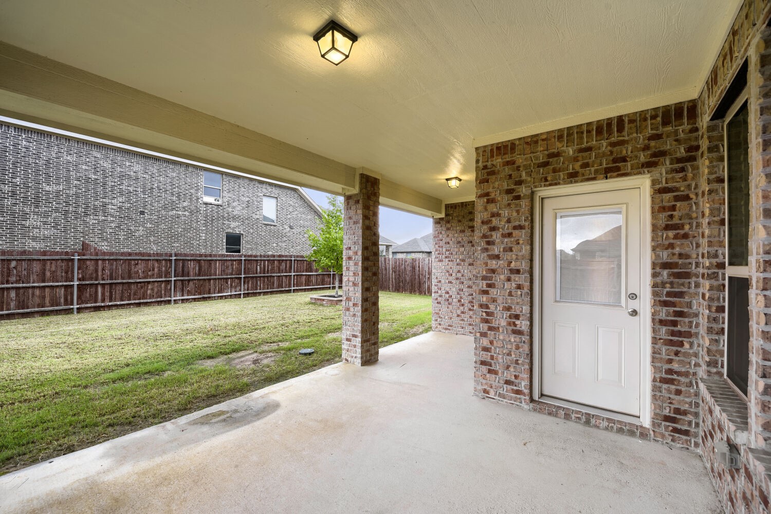 Covered concrete patio with brick walls and a white back door, overlooking a fenced grassy yard.