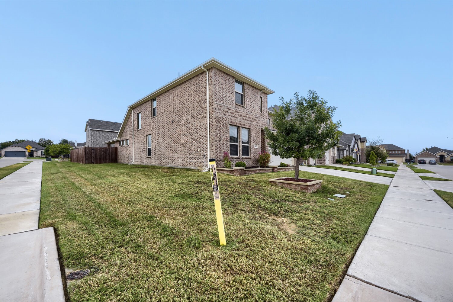 Two-story brick house on a corner lot, with a broad lawn, young tree, and sidewalks.