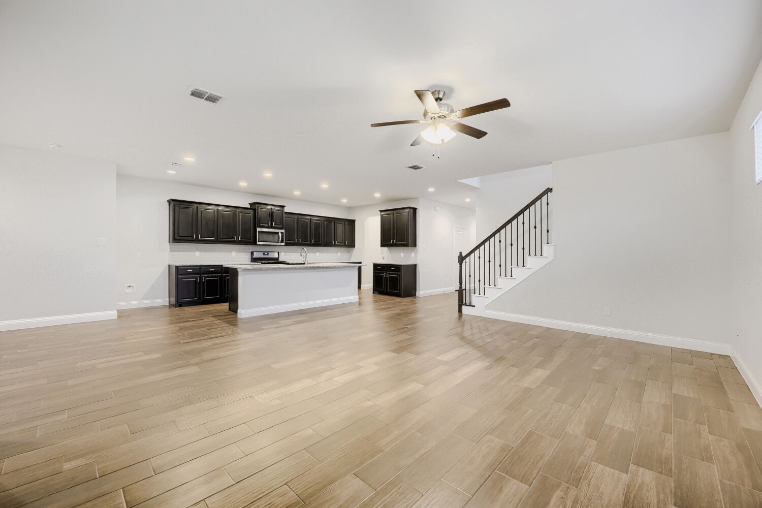 Open kitchen and living area with dark cabinets, wood-look tile flooring, and staircase.