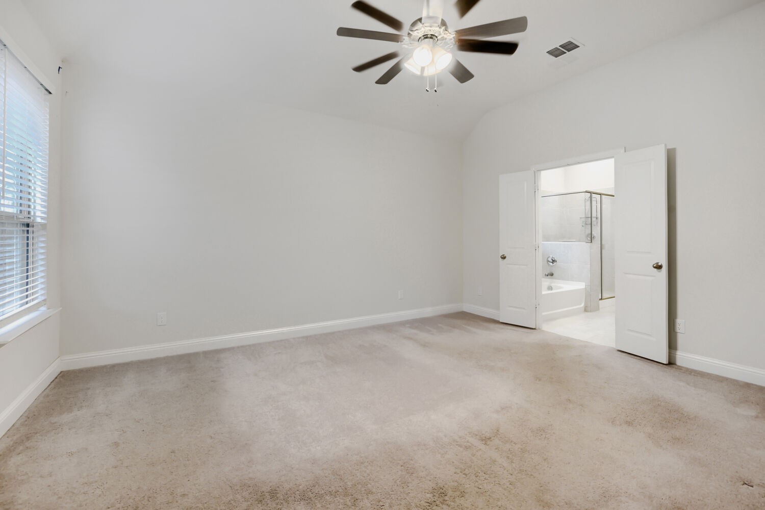 Empty carpeted bedroom with a ceiling fan, window blinds, and open doors to an ensuite bathroom.