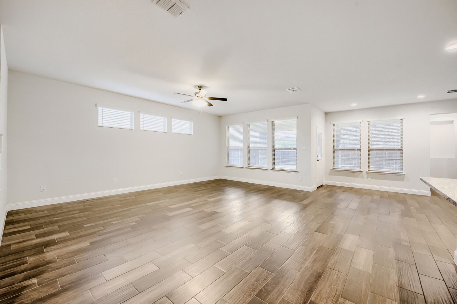 Empty living room with wood-look tile floors, multiple windows, and a ceiling fan.