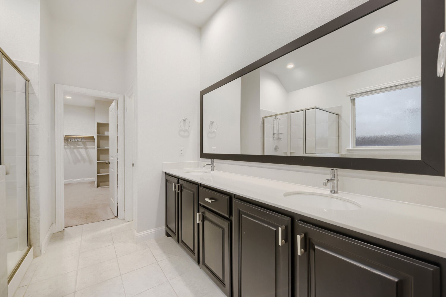 Bathroom with double-sink vanity, dark cabinetry, glass shower, and walk-in closet.