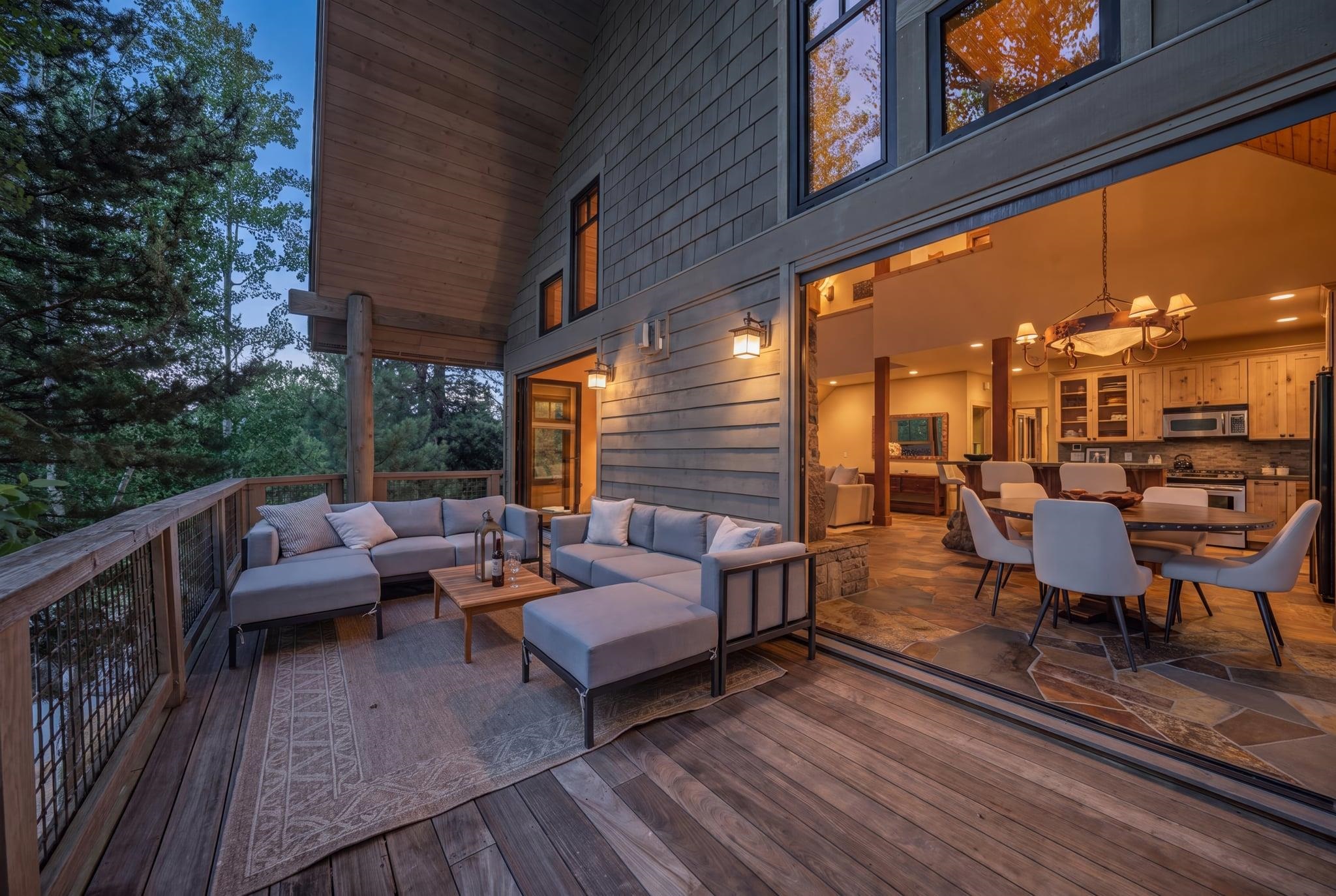 Furnished wood deck with gray lounge seating opens into a warmly lit dining room and kitchen.