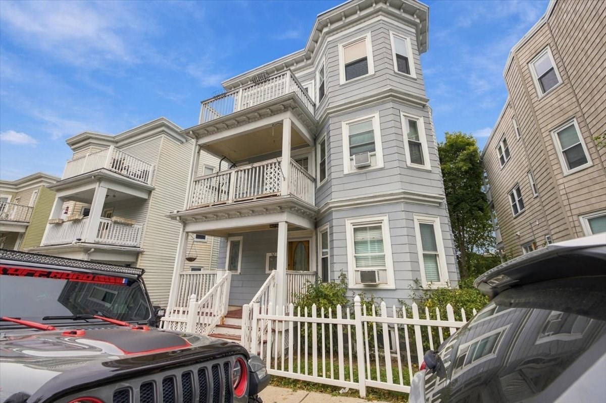 Gray multi-story house with stacked porches and bay windows behind a white picket fence.