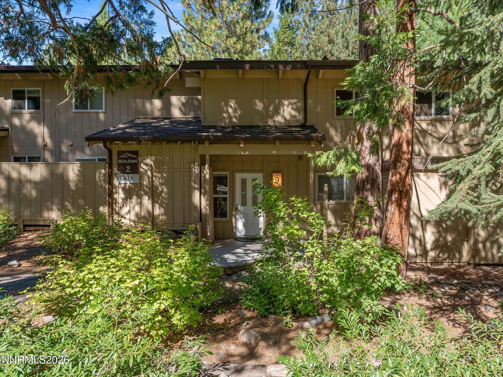 Residential building entrance framed by leafy shrubs and tall evergreen trees.