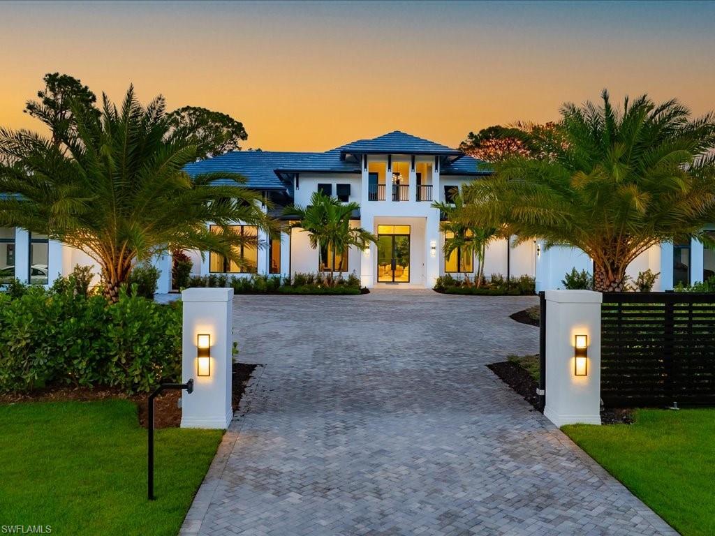 White two-story home with a blue tile roof, palm-lined driveway, and illuminated entrance at dusk.