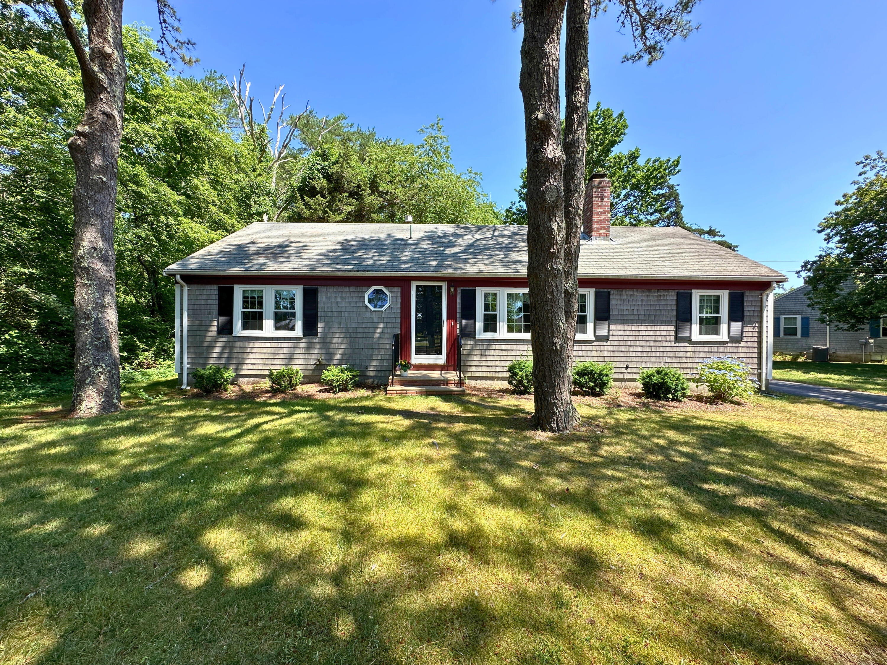 Gray shingle house with a red-trimmed entrance, framed by mature trees and a sunlit lawn.