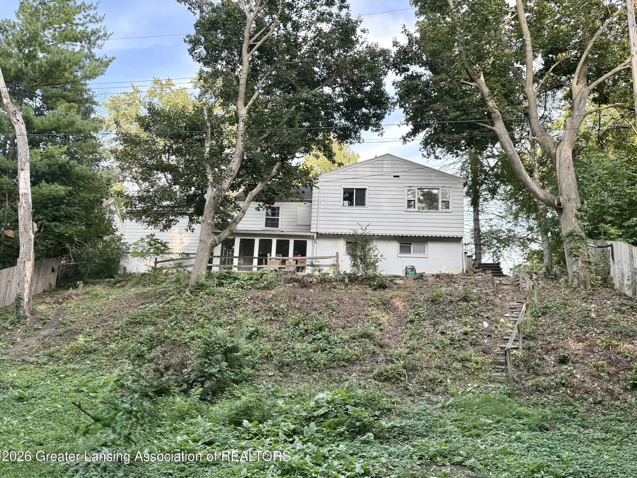 White-sided house behind mature trees, with a small deck and stone steps on a steep wooded yard.