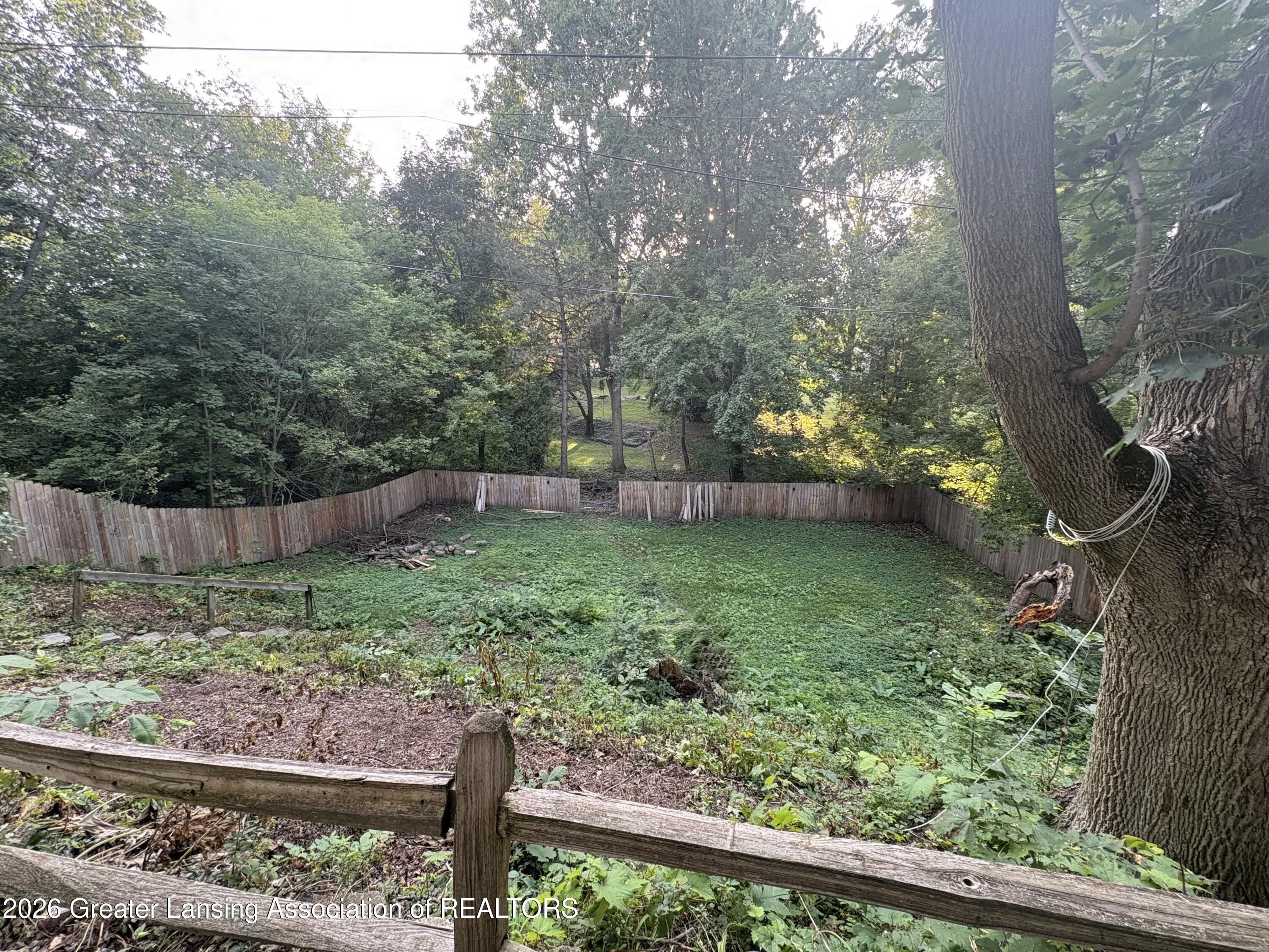 Fenced grassy backyard surrounded by mature trees, viewed over a weathered wooden rail.