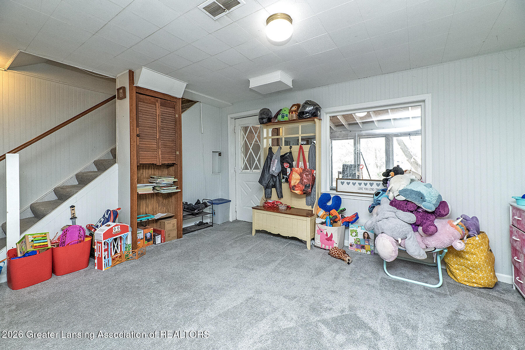 Carpeted playroom with a staircase, built-in wooden cabinet, toys, and stuffed animals.