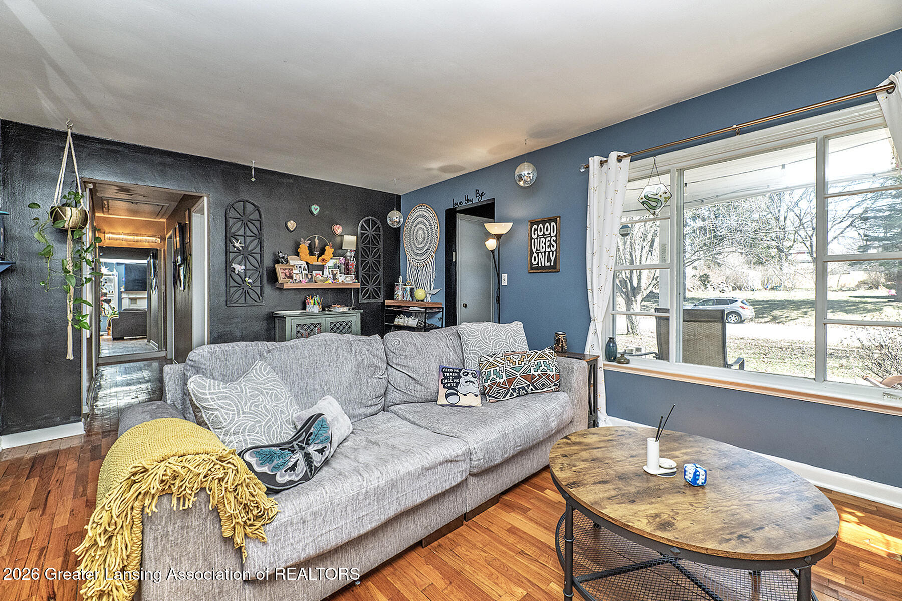 Living room with a gray sectional, yellow throw, hardwood floors, and a large window overlooking a wintry yard.