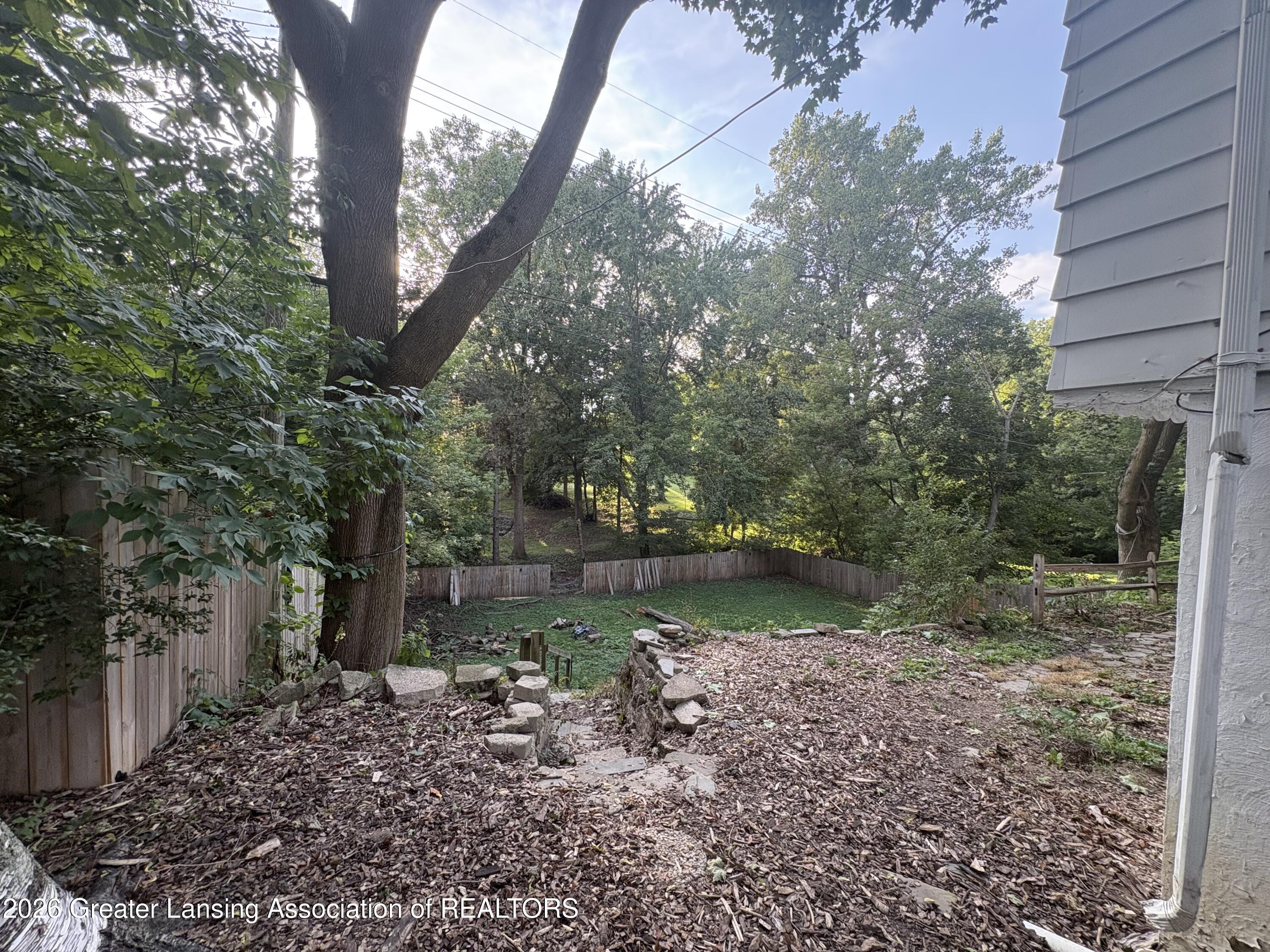 Fenced backyard with mature trees, a grassy clearing, wood-chip ground cover, and stone edging.