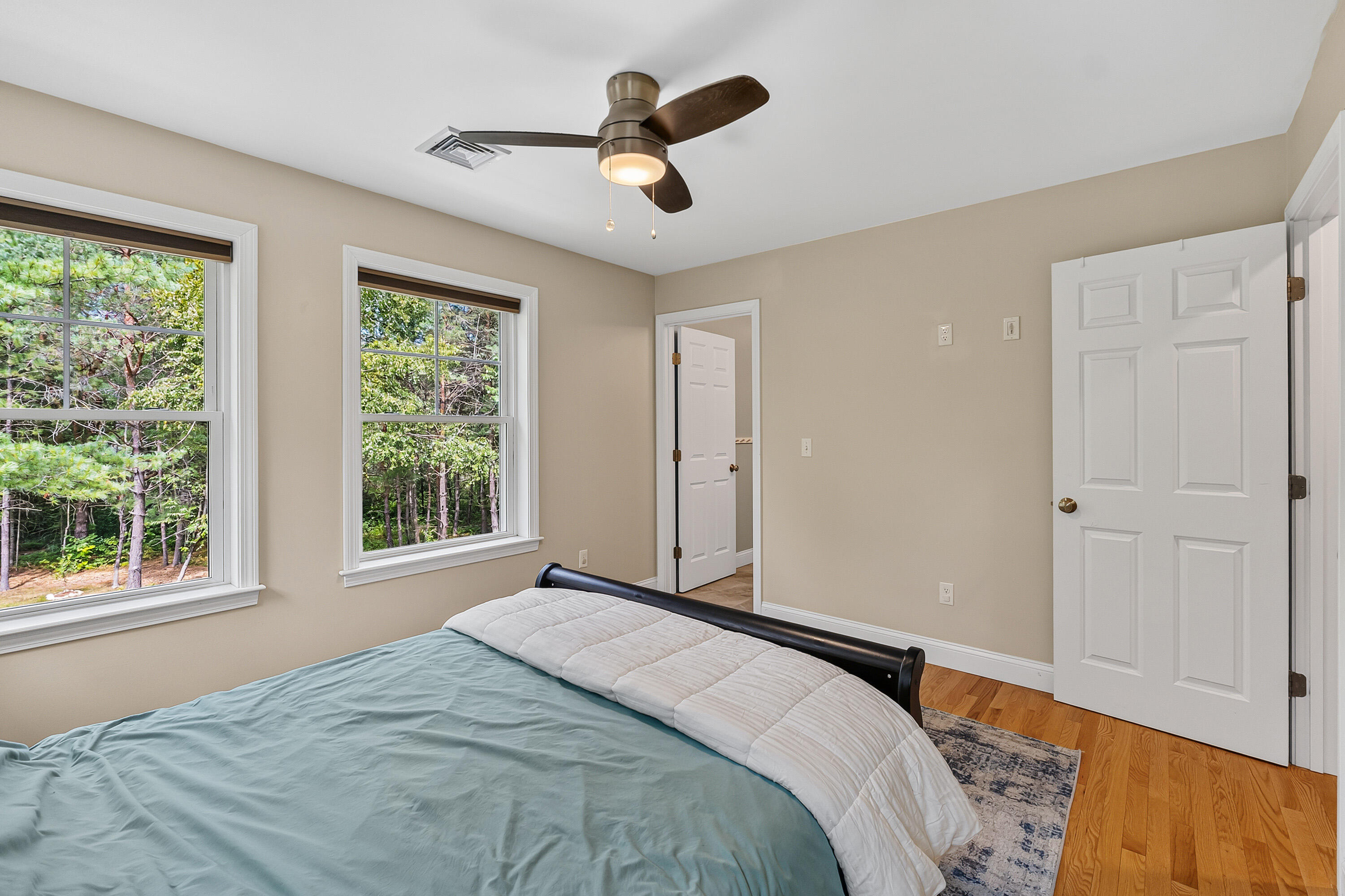 Bedroom with a bed, ceiling fan, and two windows overlooking trees.