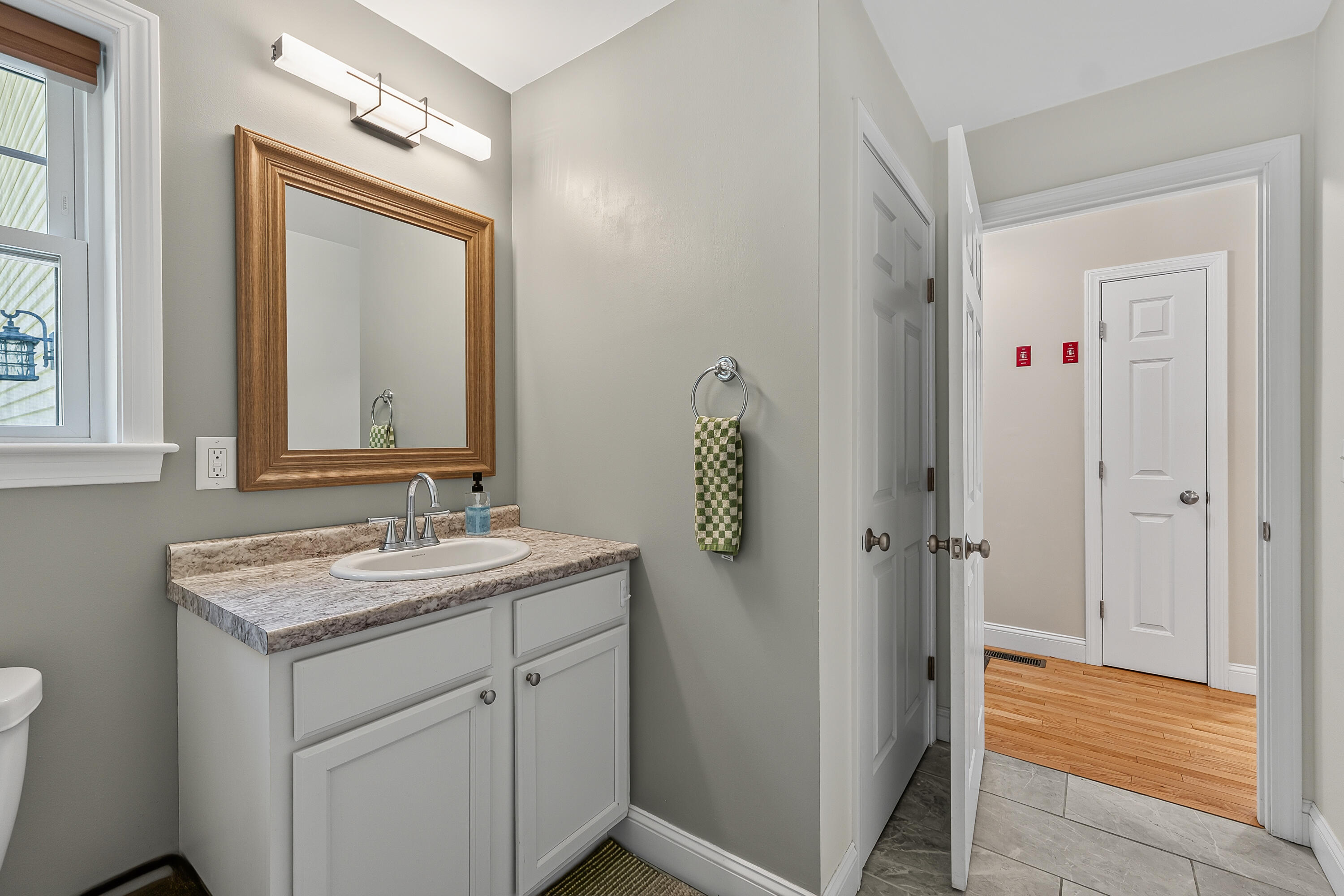 Bathroom vanity with a stone-look counter and wood-framed mirror beside an open doorway to a hardwood hall.