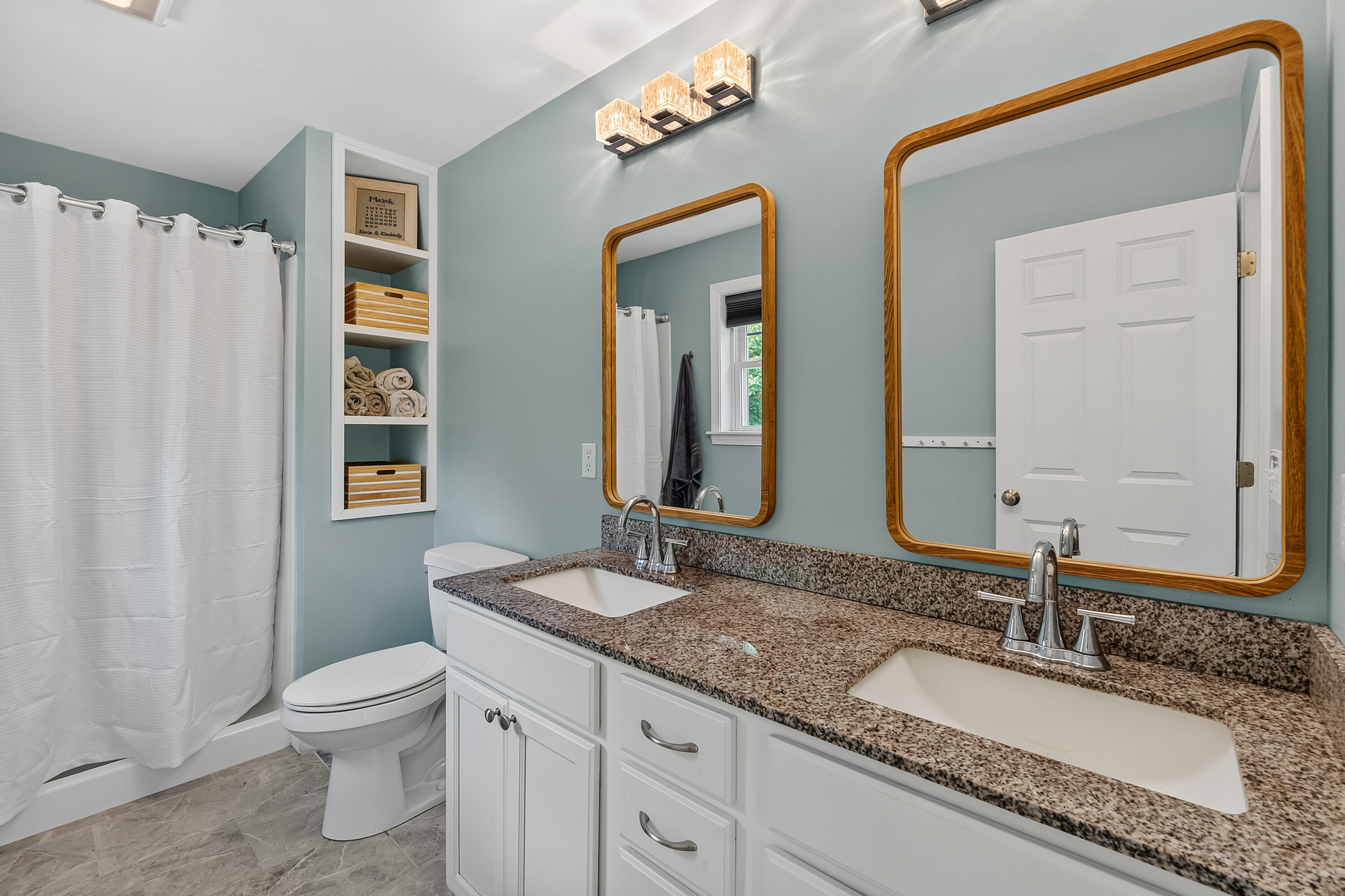 Bathroom with a double-sink granite vanity, wood-framed mirrors, built-in shelves, and a white shower curtain.