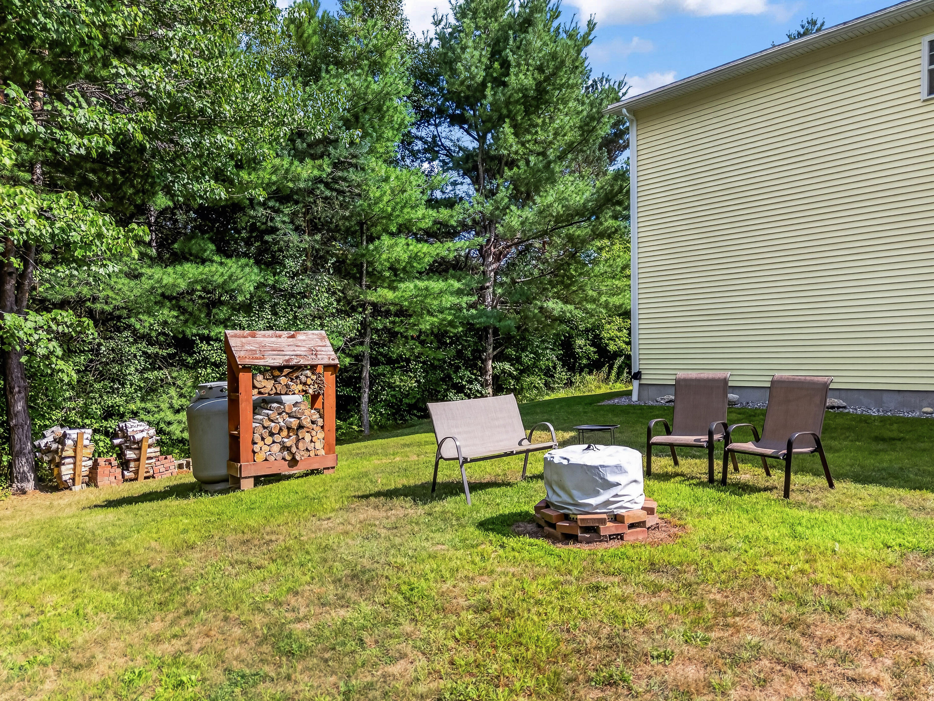 Backyard with outdoor chairs and a covered fire pit beside a wooded lawn.